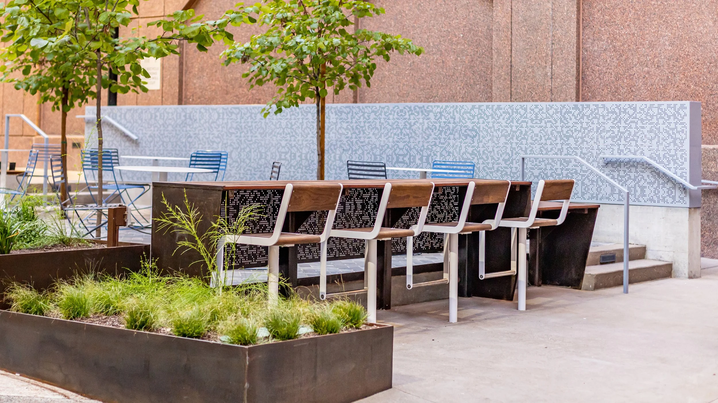 Outdoor seating area with a wooden bar counter, modern chairs, small trees, and planters with green plants, against a textured wall with stairs in the background.