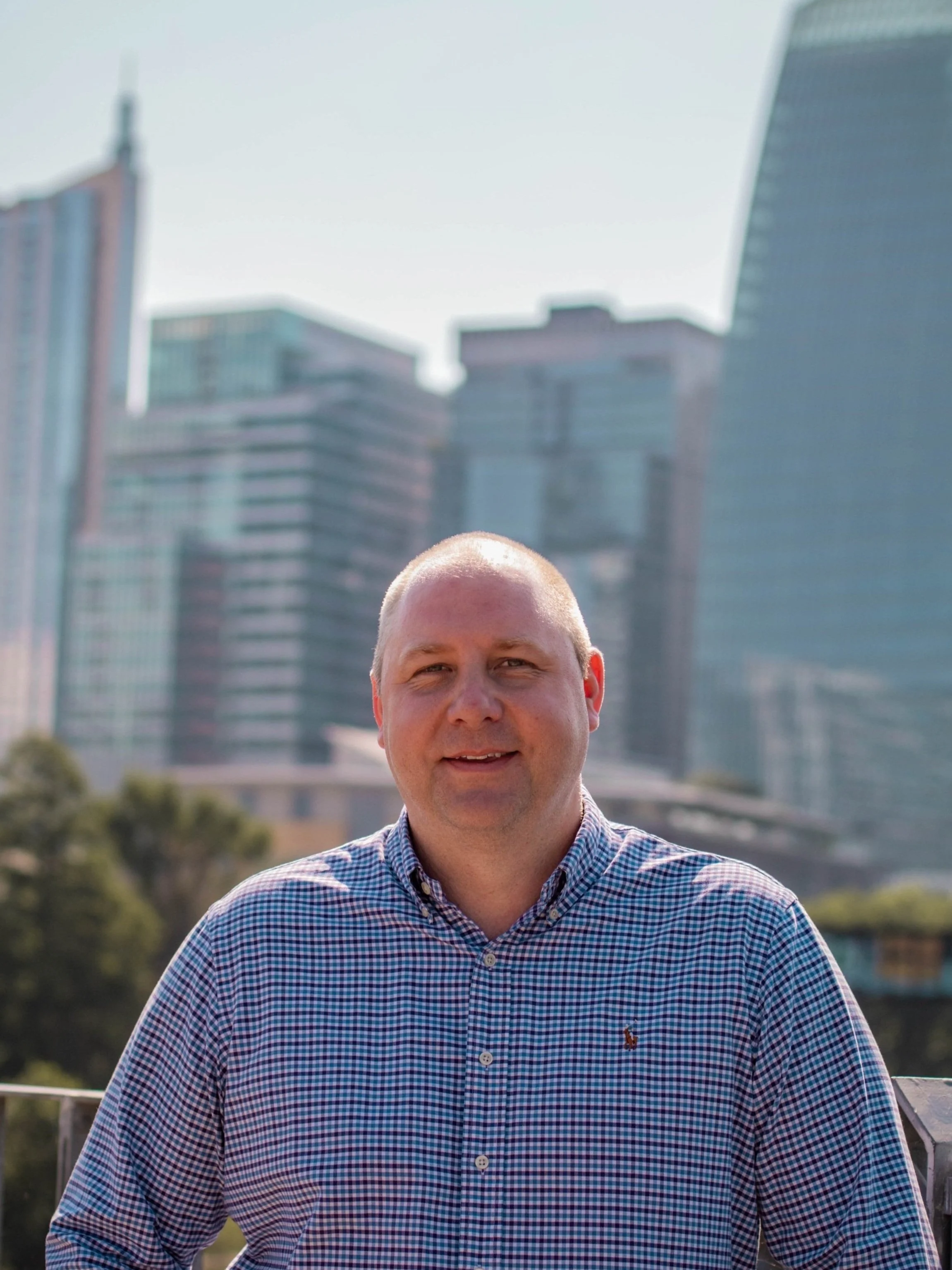 Man with short blonde hair wearing a checkered shirt outdoors with city skyscrapers in the background.