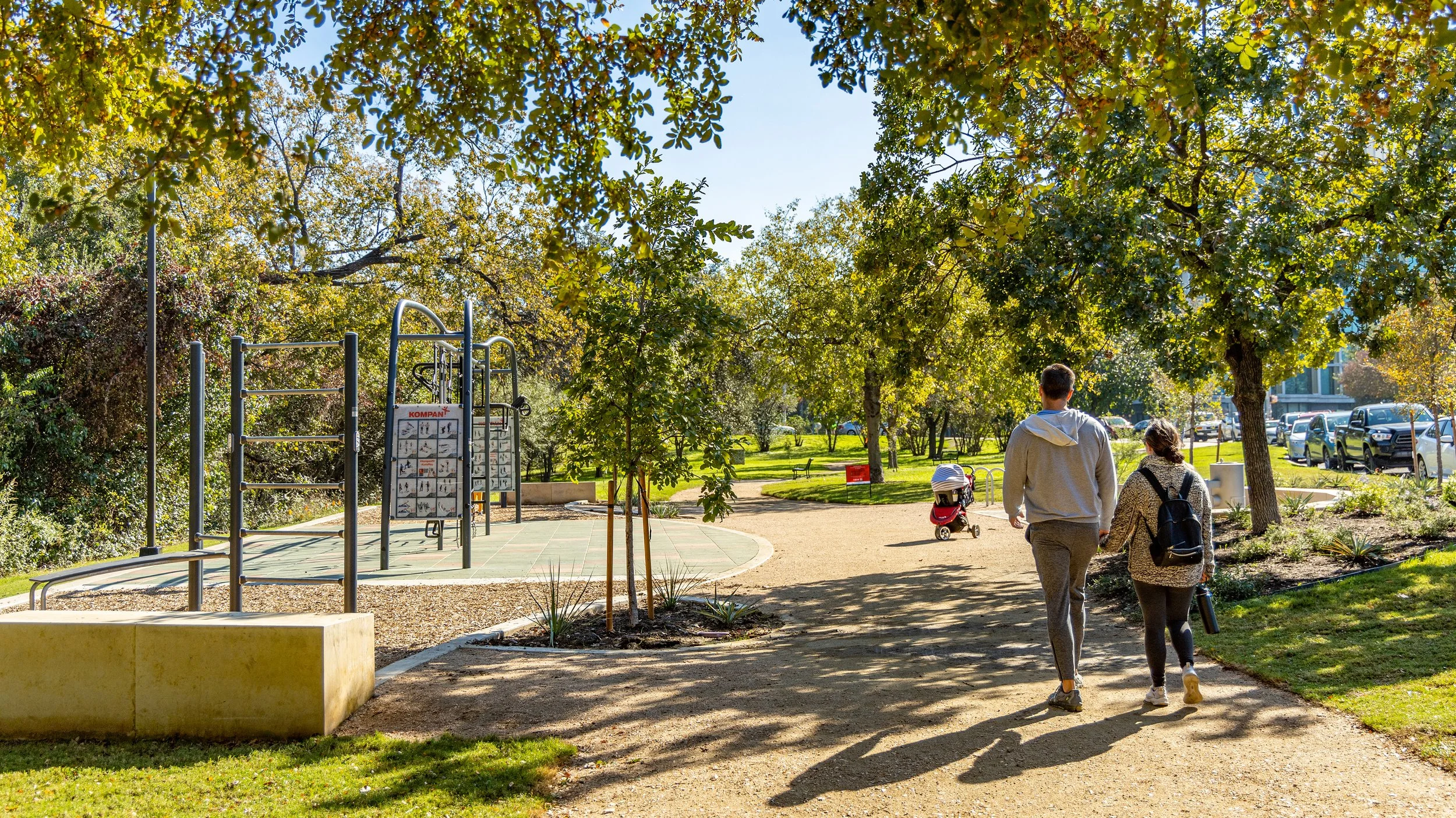 Two people walking on a shaded park path, surrounded by trees and greenery, with a playground on the left and parked cars on the right.