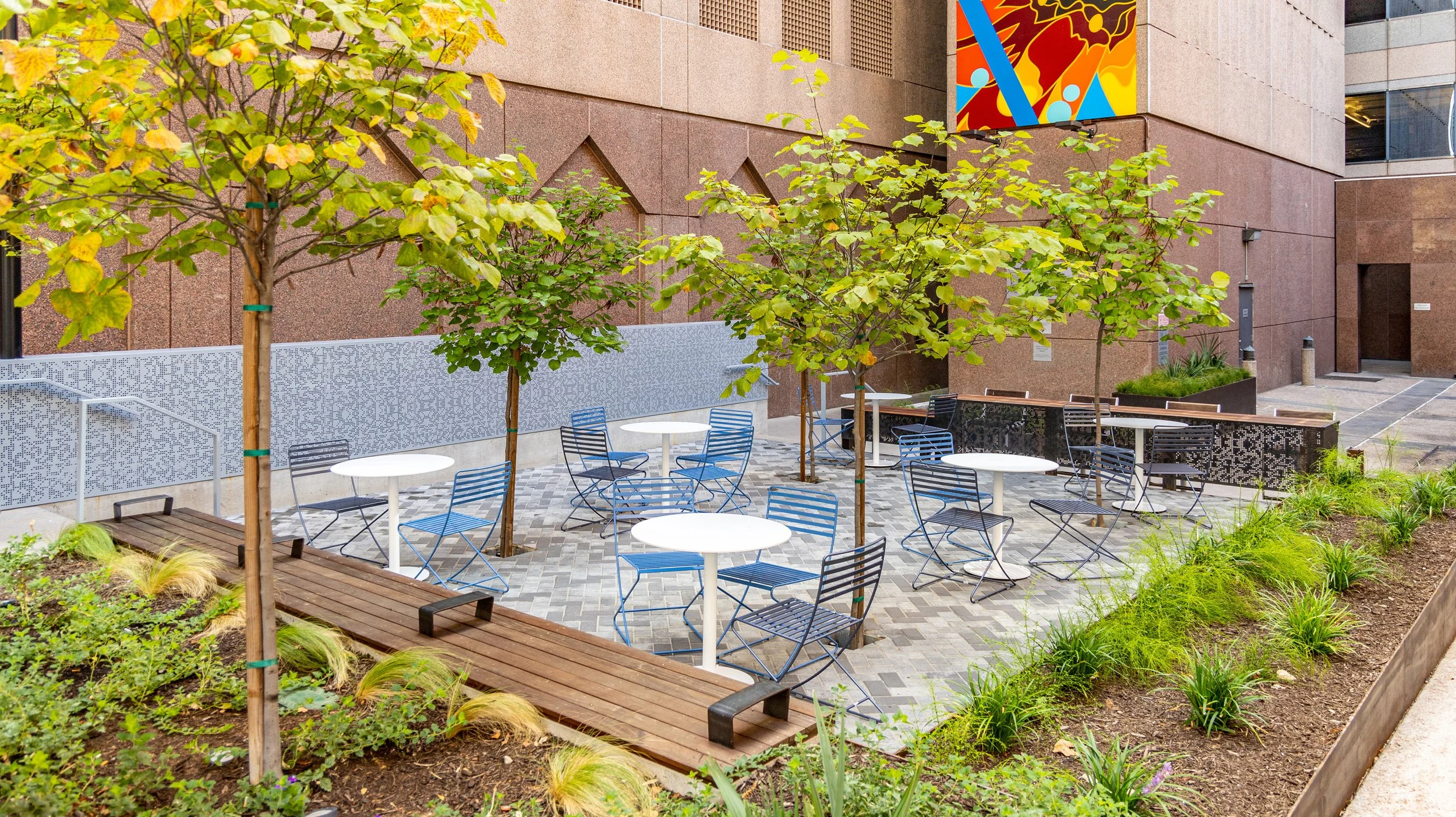 Outdoor seating area with blue chairs and white tables, surrounded by small trees and greenery, in an urban setting.