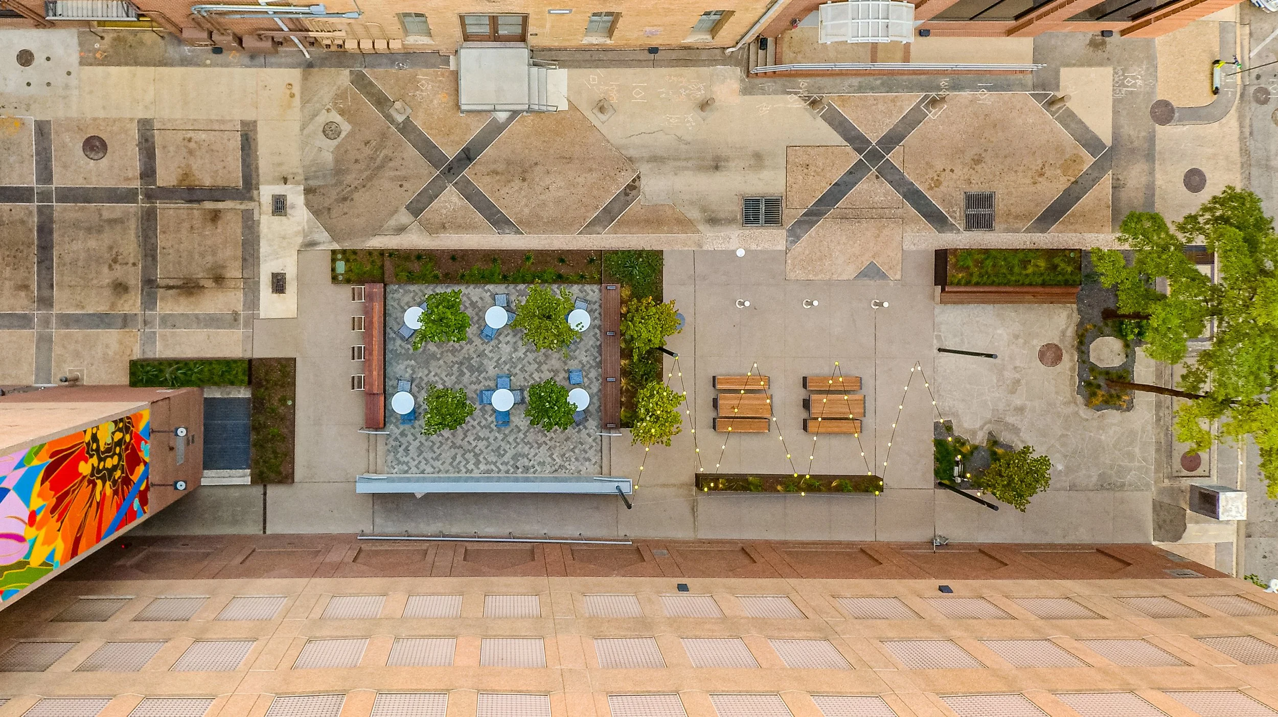 Aerial view of an urban outdoor courtyard featuring small round tables with chairs, benches, string lights, trees, and surrounding buildings.
