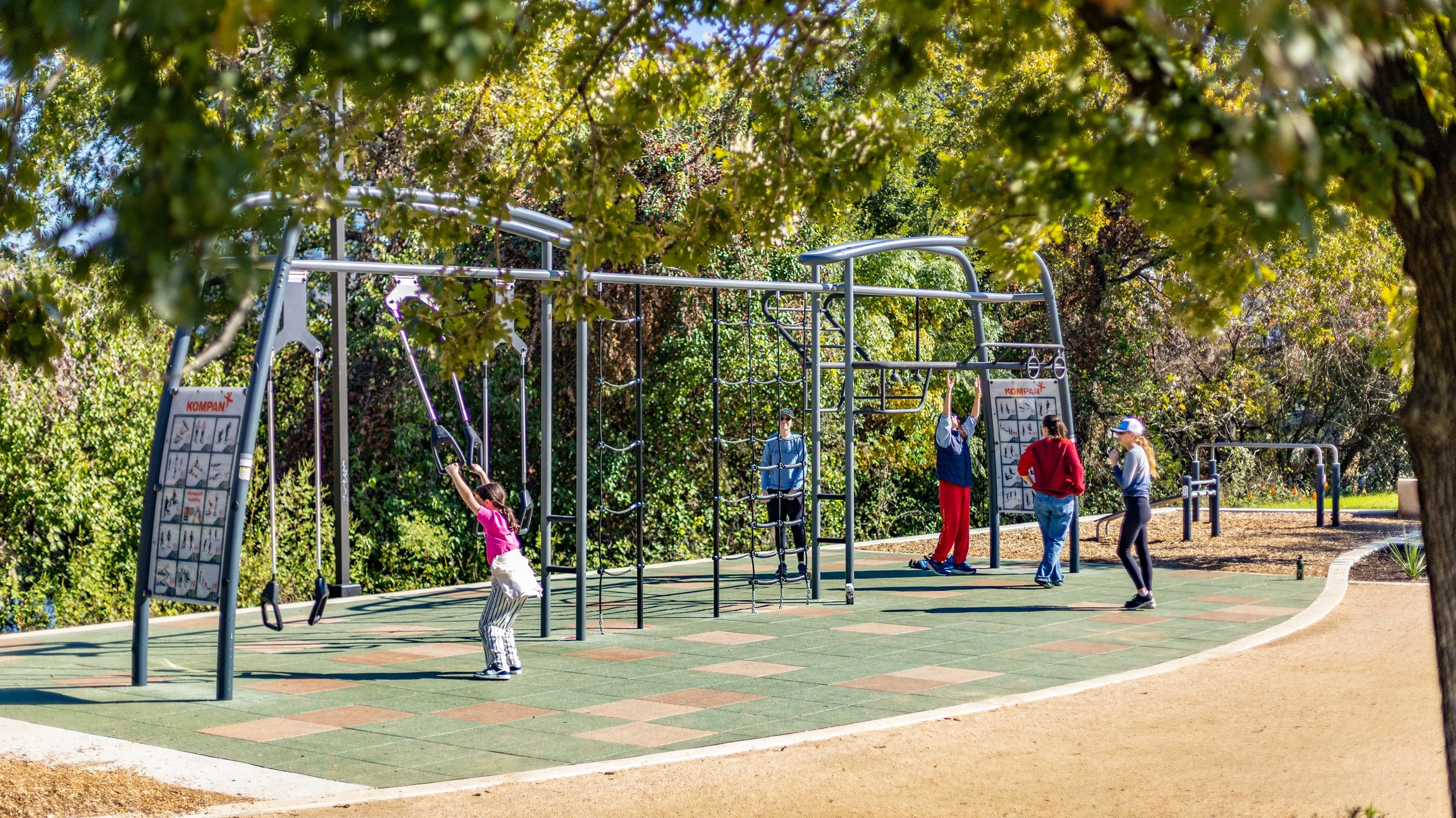 Children and adults playing and exercising on a playground with climbing equipment, swings, and fitness boards, surrounded by trees and greenery.