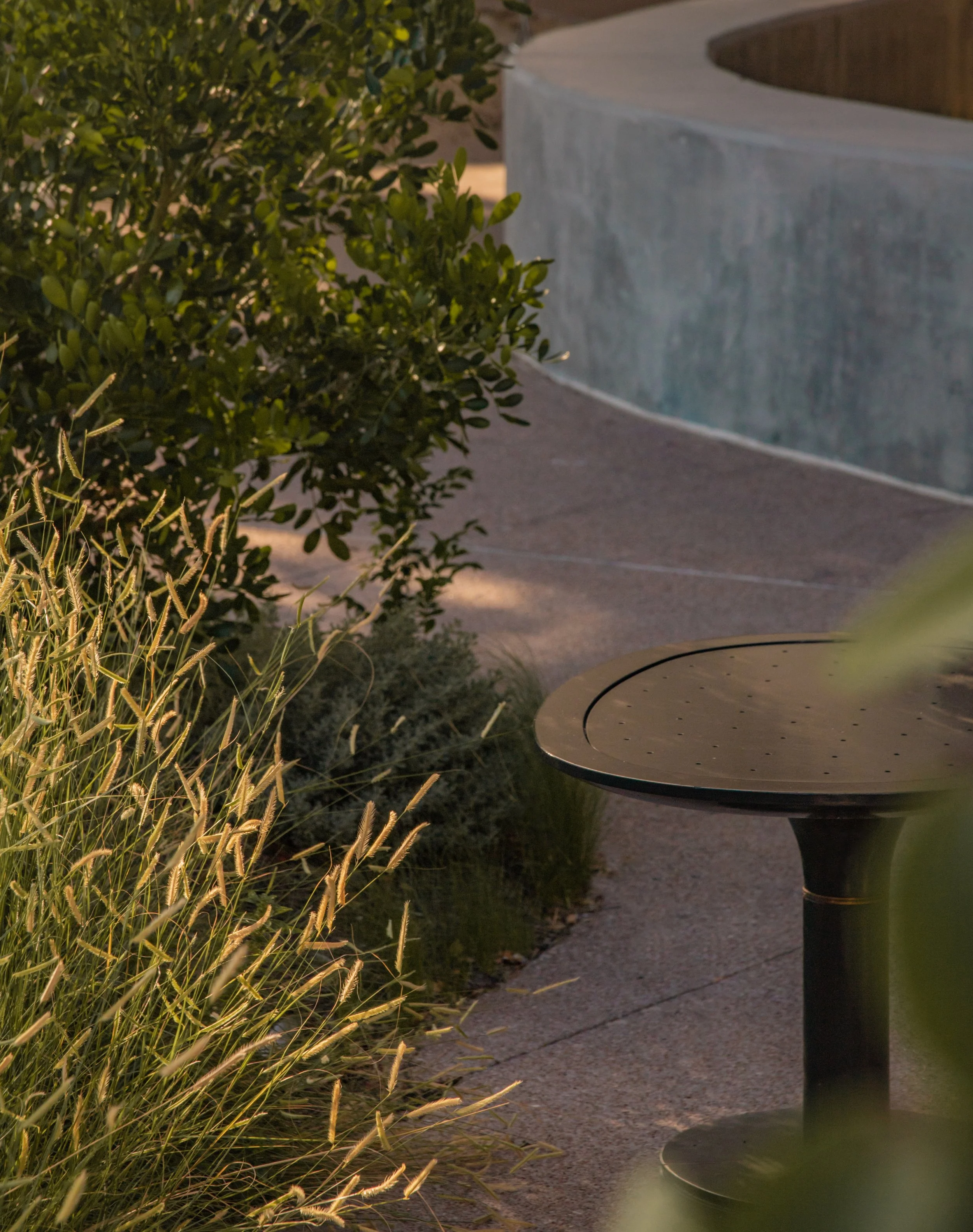 A black outdoor table with a round, smooth surface near a sidewalk, surrounded by green bushes and plants, with a concrete structure in the background.