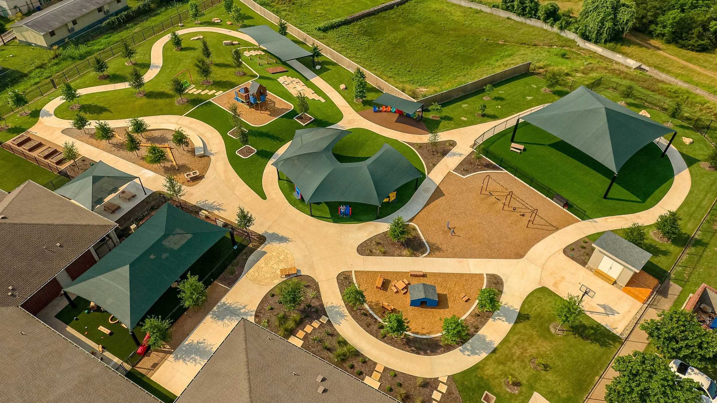 Aerial view of a school playground with walking paths, covered pavilions, playground equipment, benches, trees, and landscaped areas.