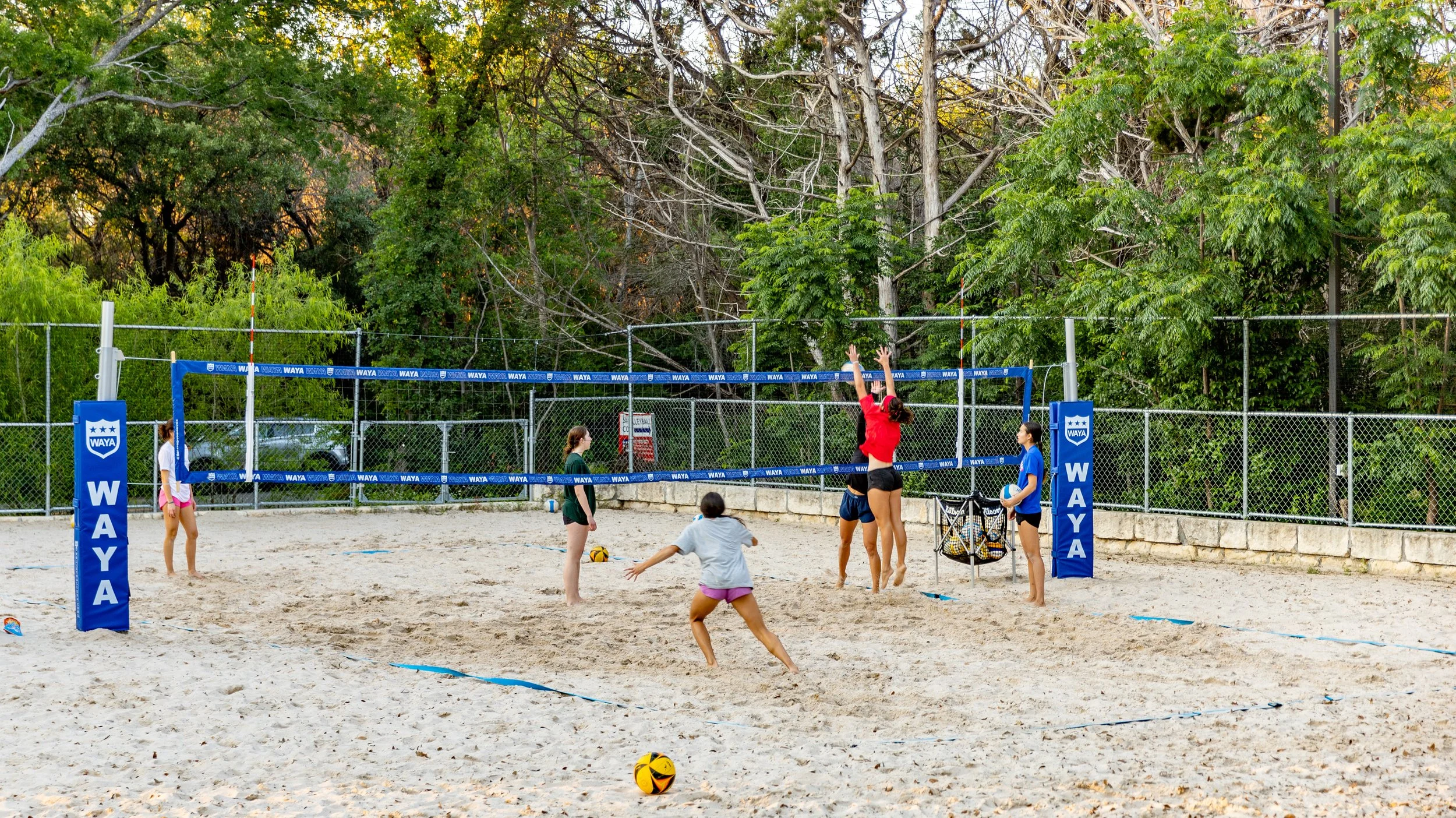Children playing volleyball on a sandy court surrounded by trees.