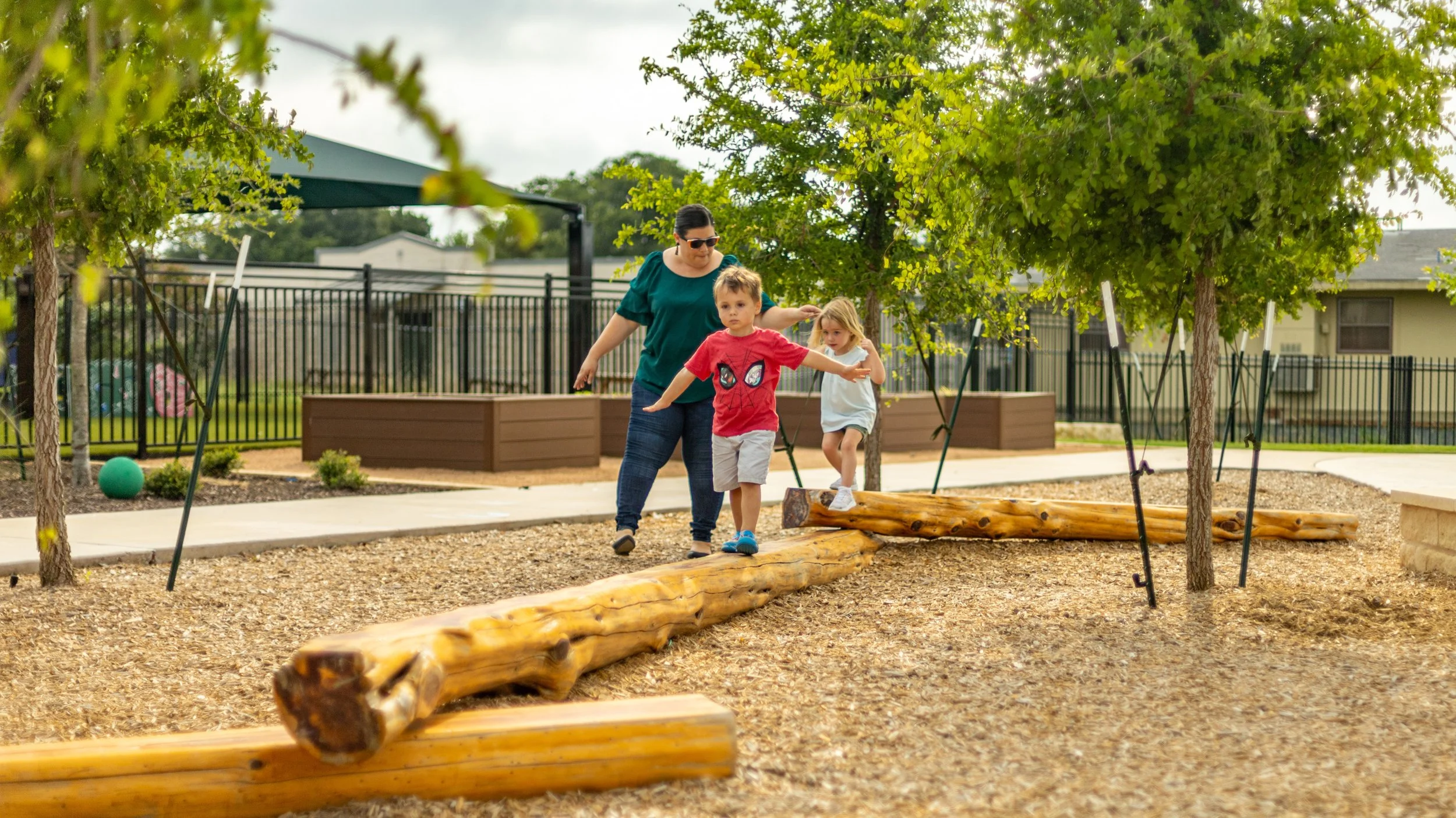 A woman helping two children walk on a wooden balance beam in a playground with trees, mulch, and a fence in the background.