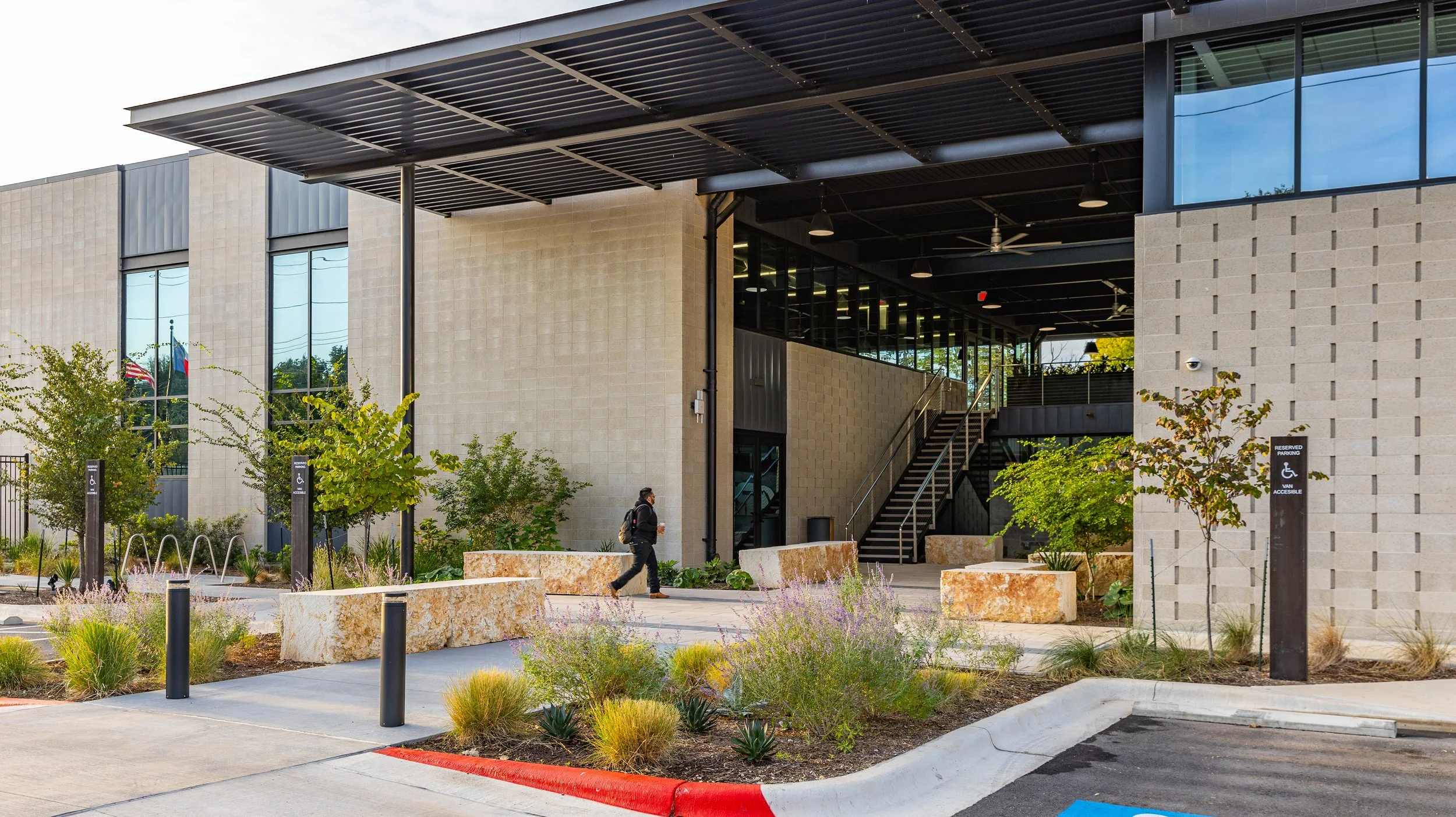 Exterior view of a modern building with large glass windows, an outdoor staircase, landscaped greenery, and a parking lot with reserved handicapped parking signs.
