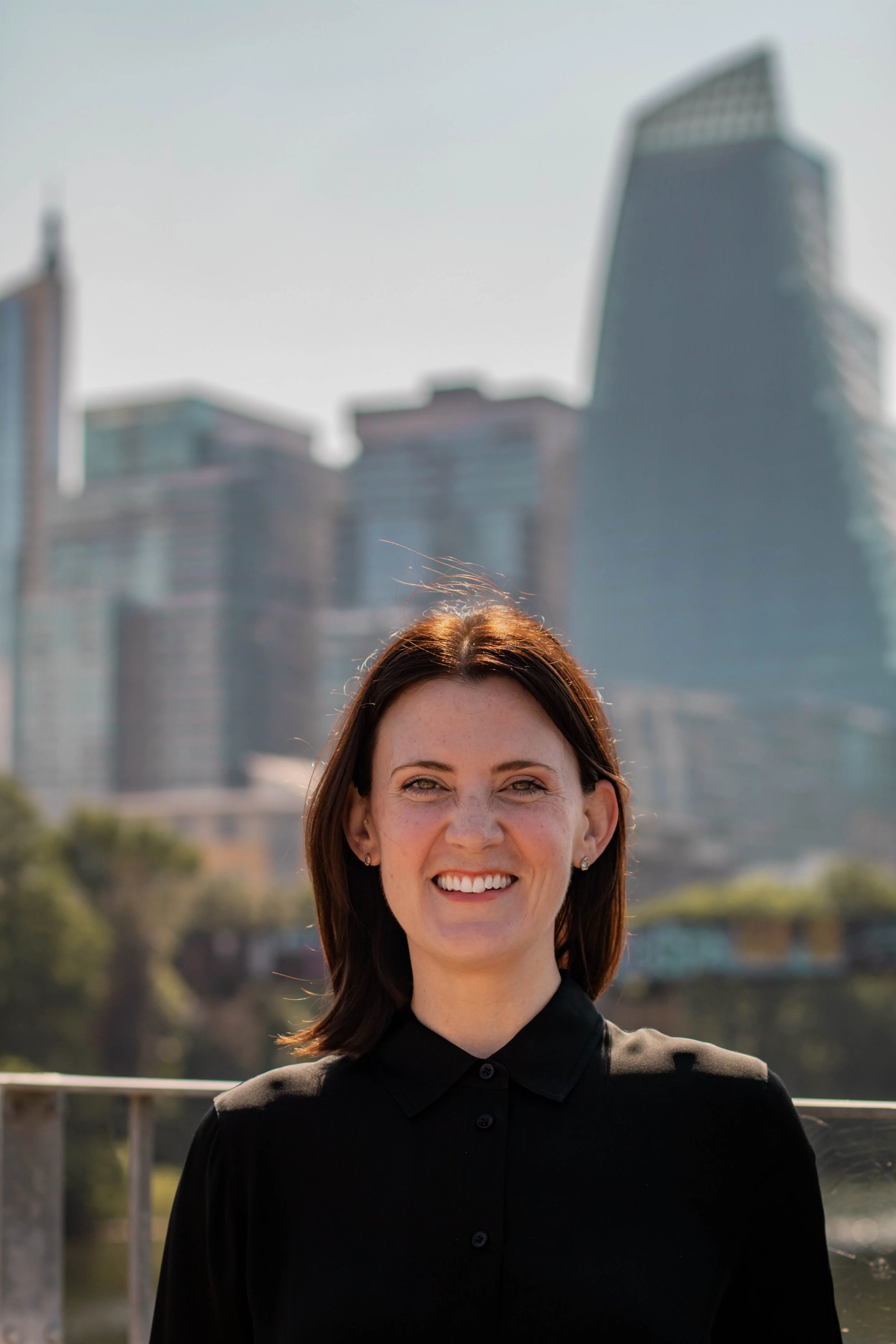 A woman with shoulder-length brown hair smiling outdoors in front of a city skyline with tall buildings in the background.