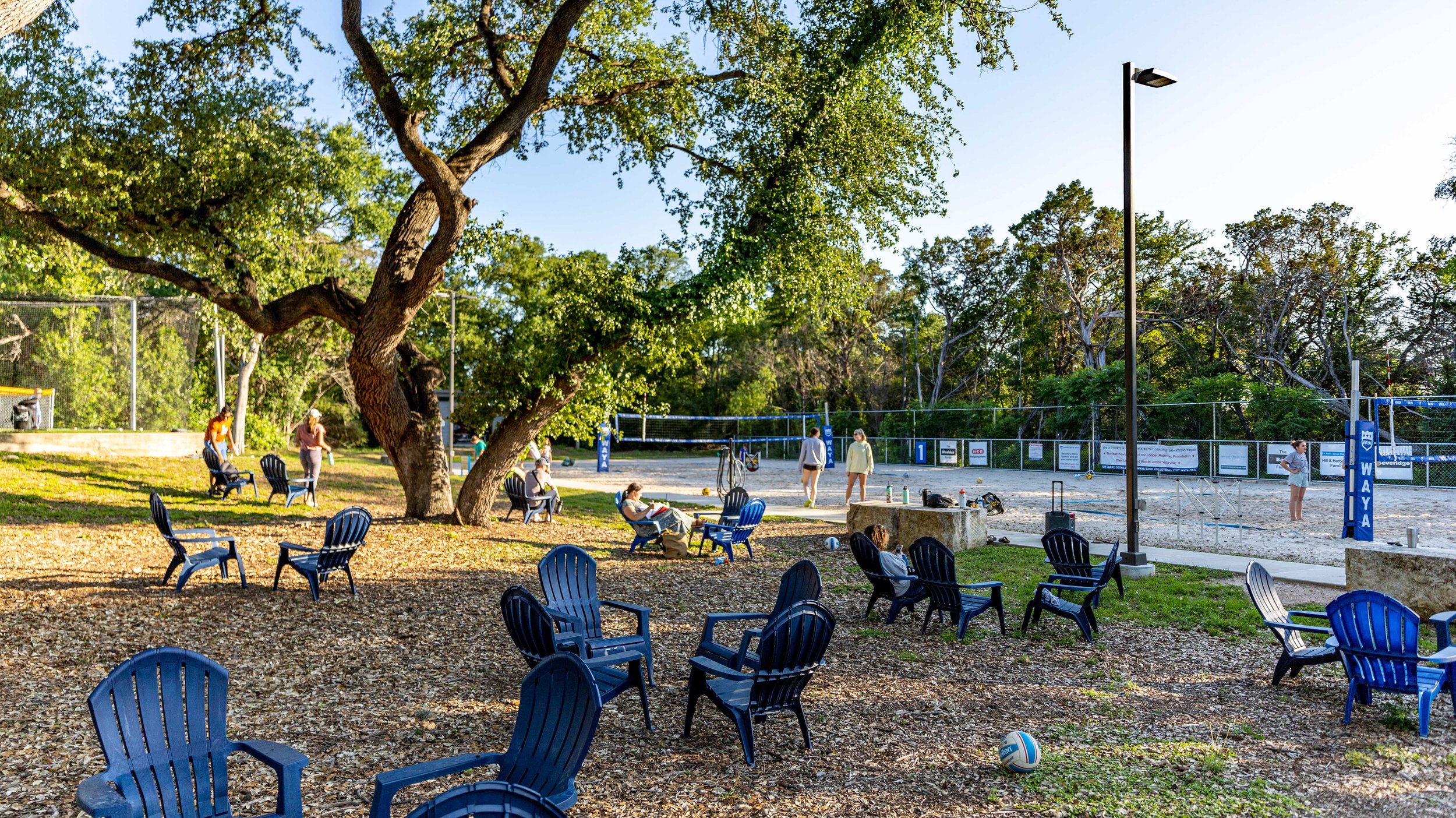 A park with a sand volleyball court, surrounded by trees and benches. Several people are playing, sitting, and relaxing, with a volleyball on the ground in front of the benches.