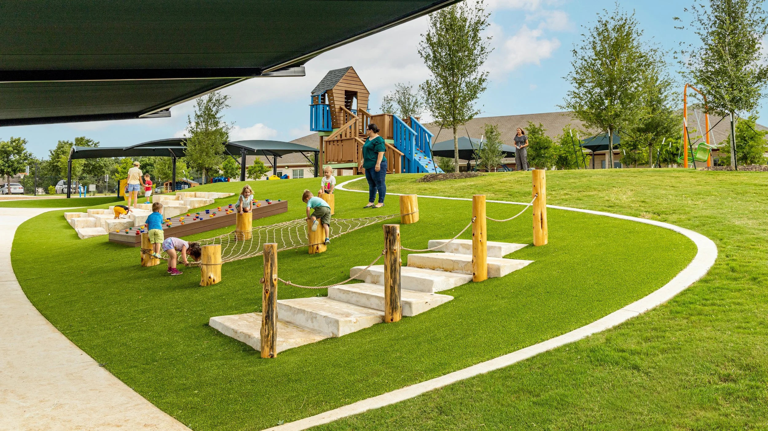Children and adults playing on a grassy playground with a small wooden castle and a climbing net, surrounded by trees and a clear sky.