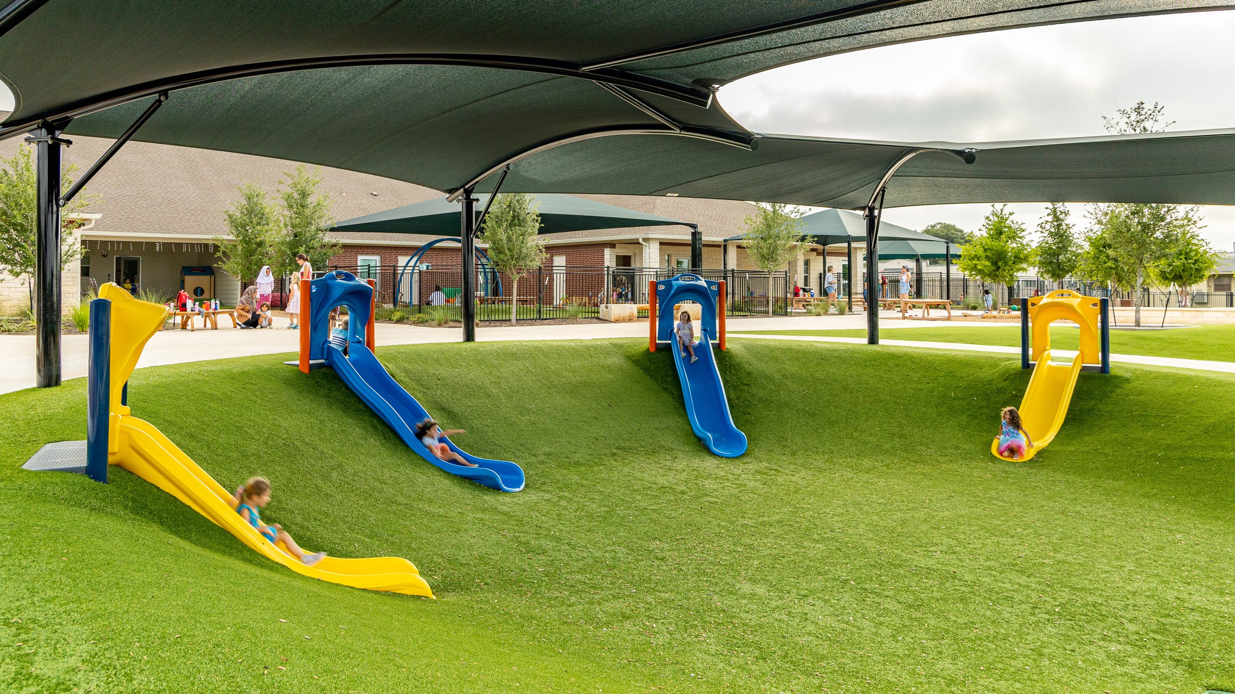 Children playing on colorful slides in a shaded outdoor playground area with trees, benches, and adults nearby.