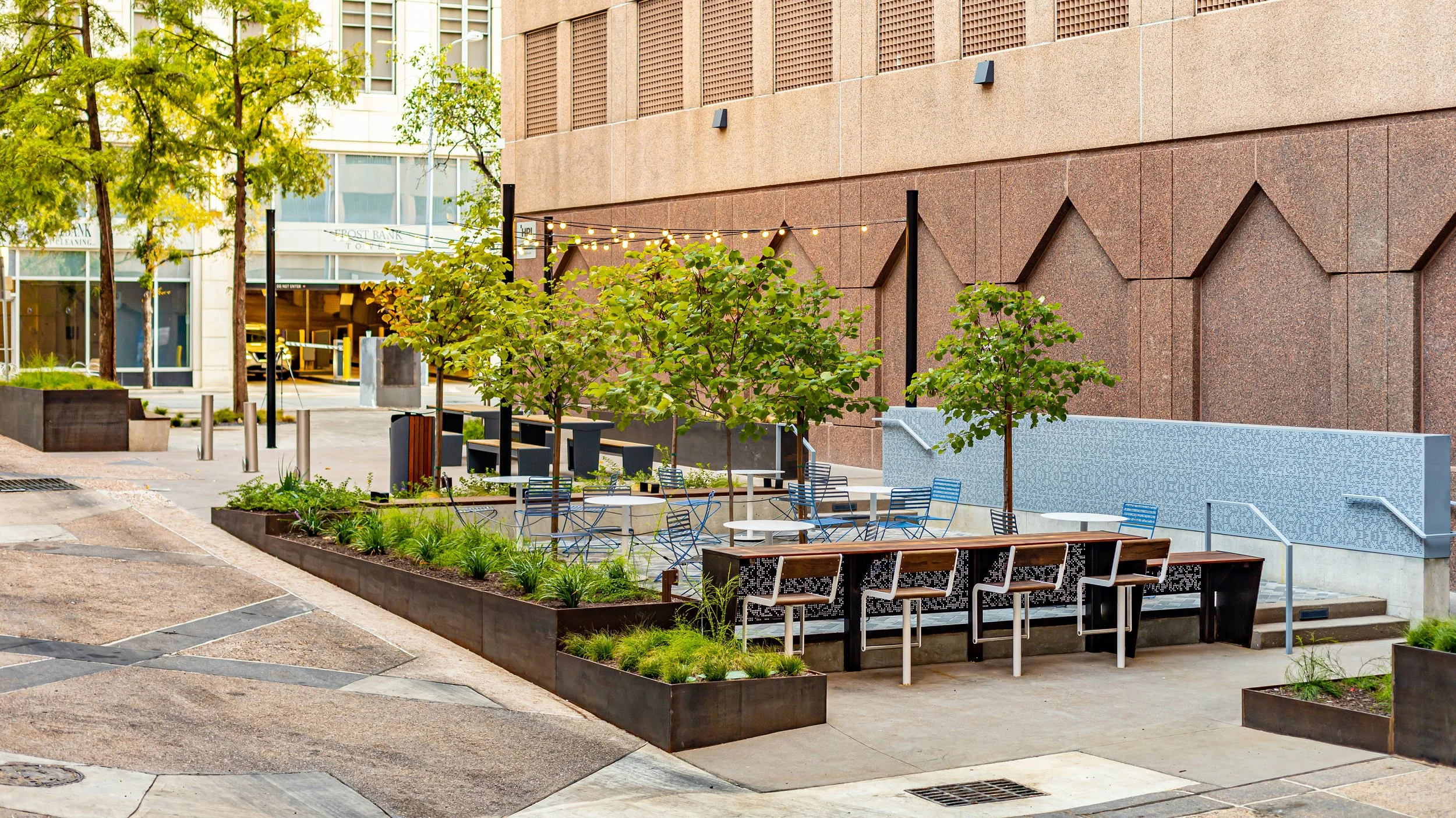 Outdoor patio with small trees, metal chairs, tables, string lights, and landscaped planters next to a red brick building.