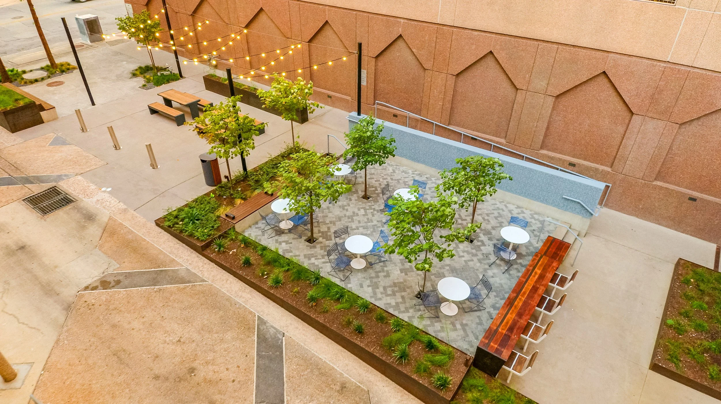 An outdoor seating area with small round tables and chairs, surrounded by trees and landscaped plants, with string lights overhead and a brick wall in the background.