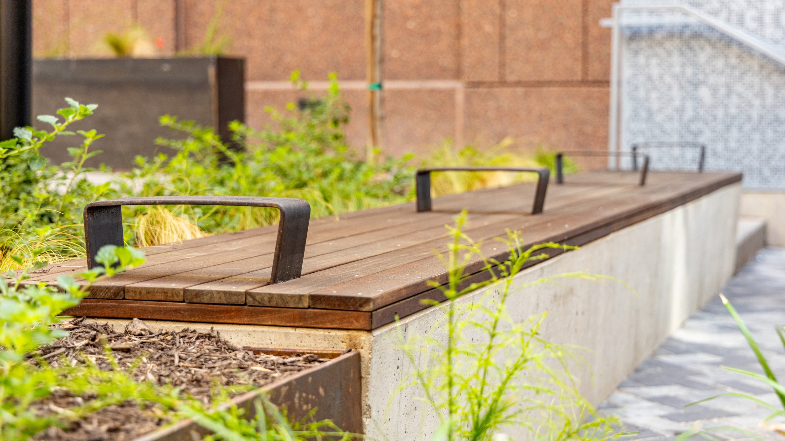 Wooden bench with black metal handles in a garden with green plants and a brick wall background.