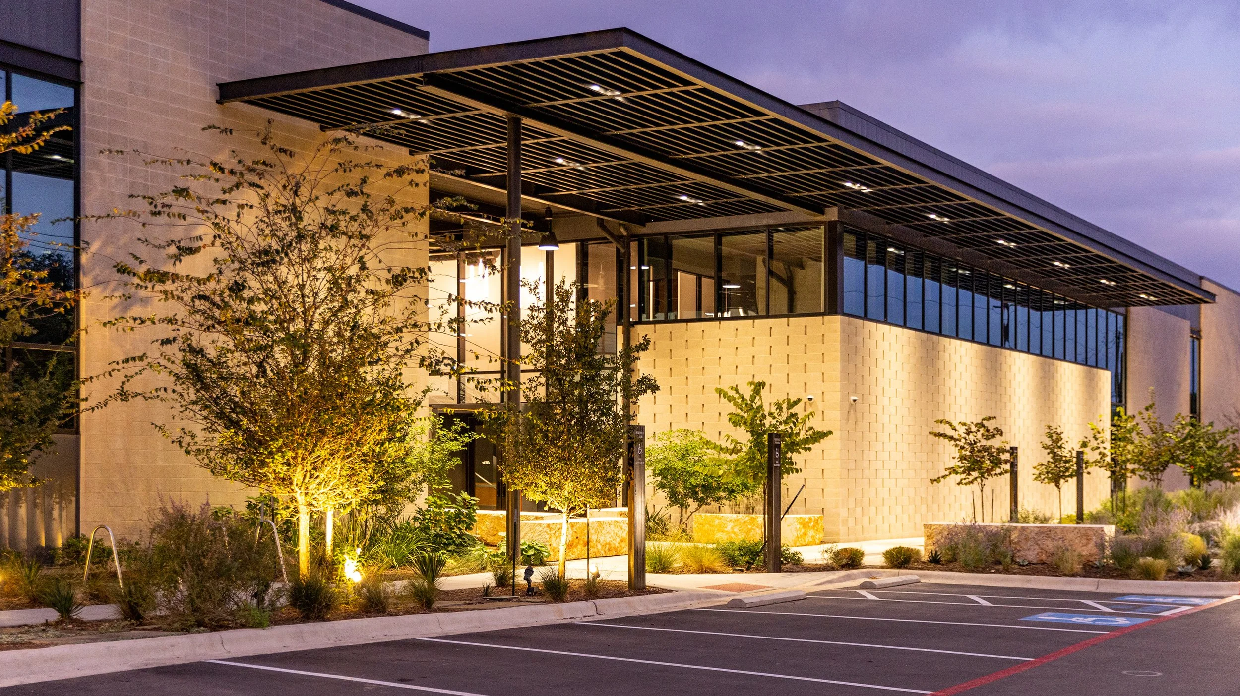 Modern office building with glass windows and landscaped parking lot at dusk, featuring trees, shrubs, and outdoor lighting.