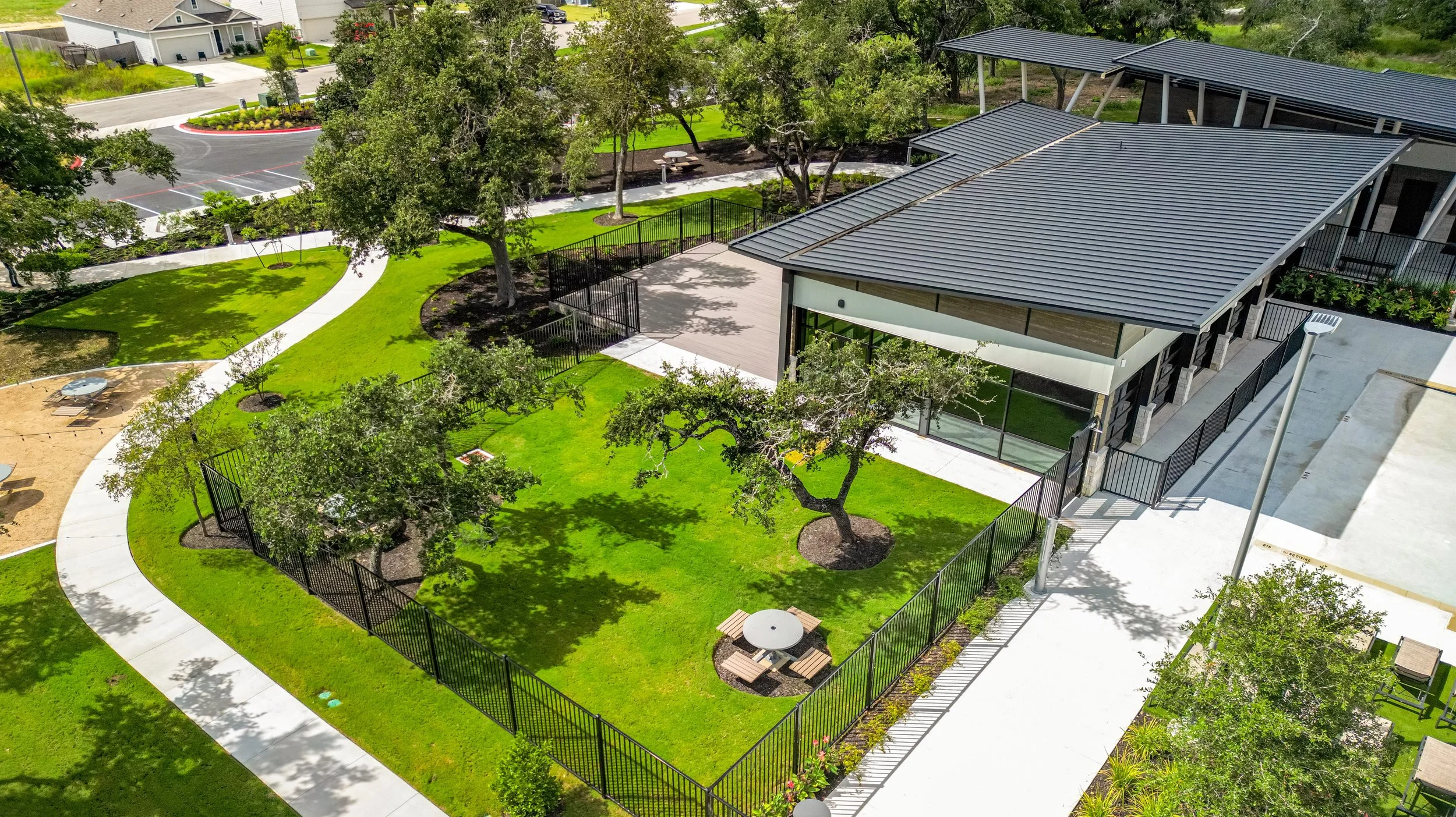 A landscaped outdoor area with a building, trees, grassy lawn, walking paths, and outdoor seating, surrounded by a black metal fence.