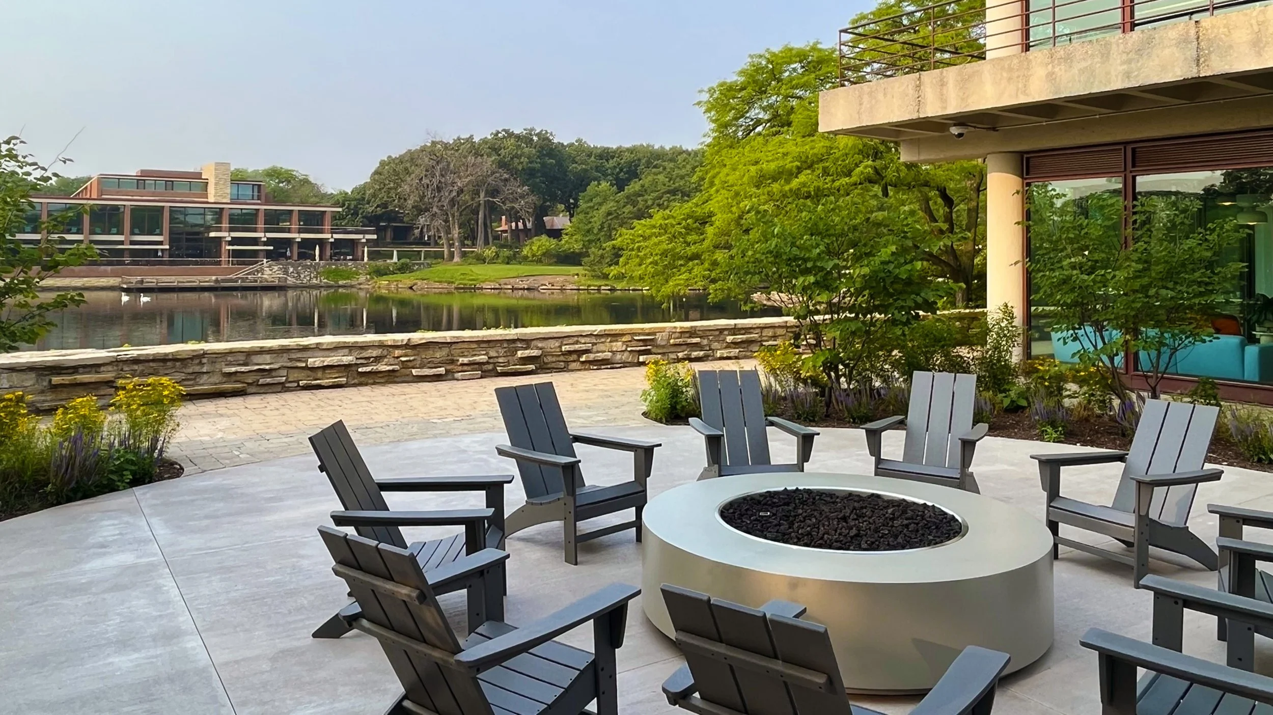 Outdoor patio with nearby water, modern building with glass windows, trees, and a garden with plants and flowers.