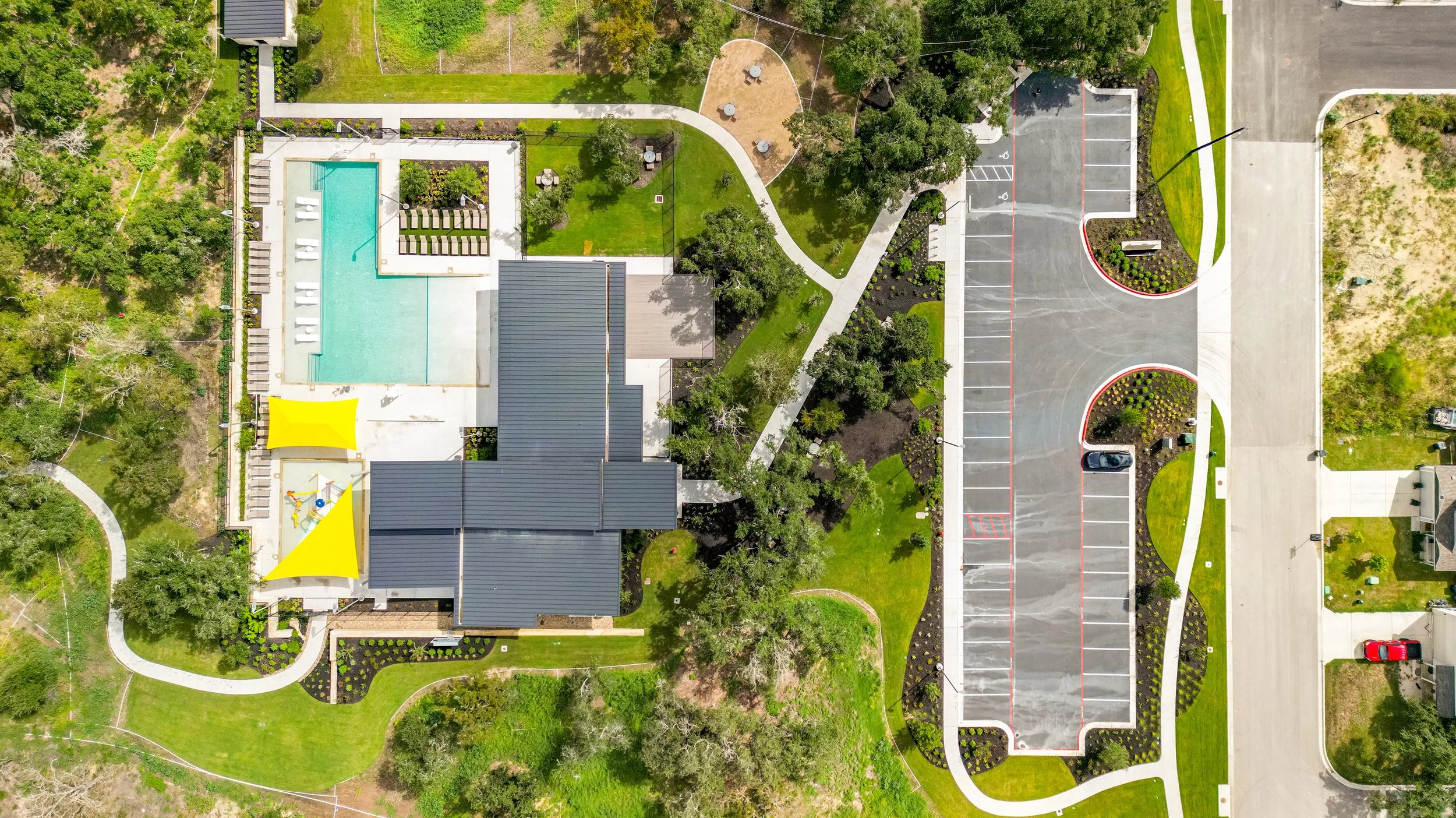 Aerial view of a building with swimming pool, yellow shade structures, playground, and surrounding greenery and parking lot.