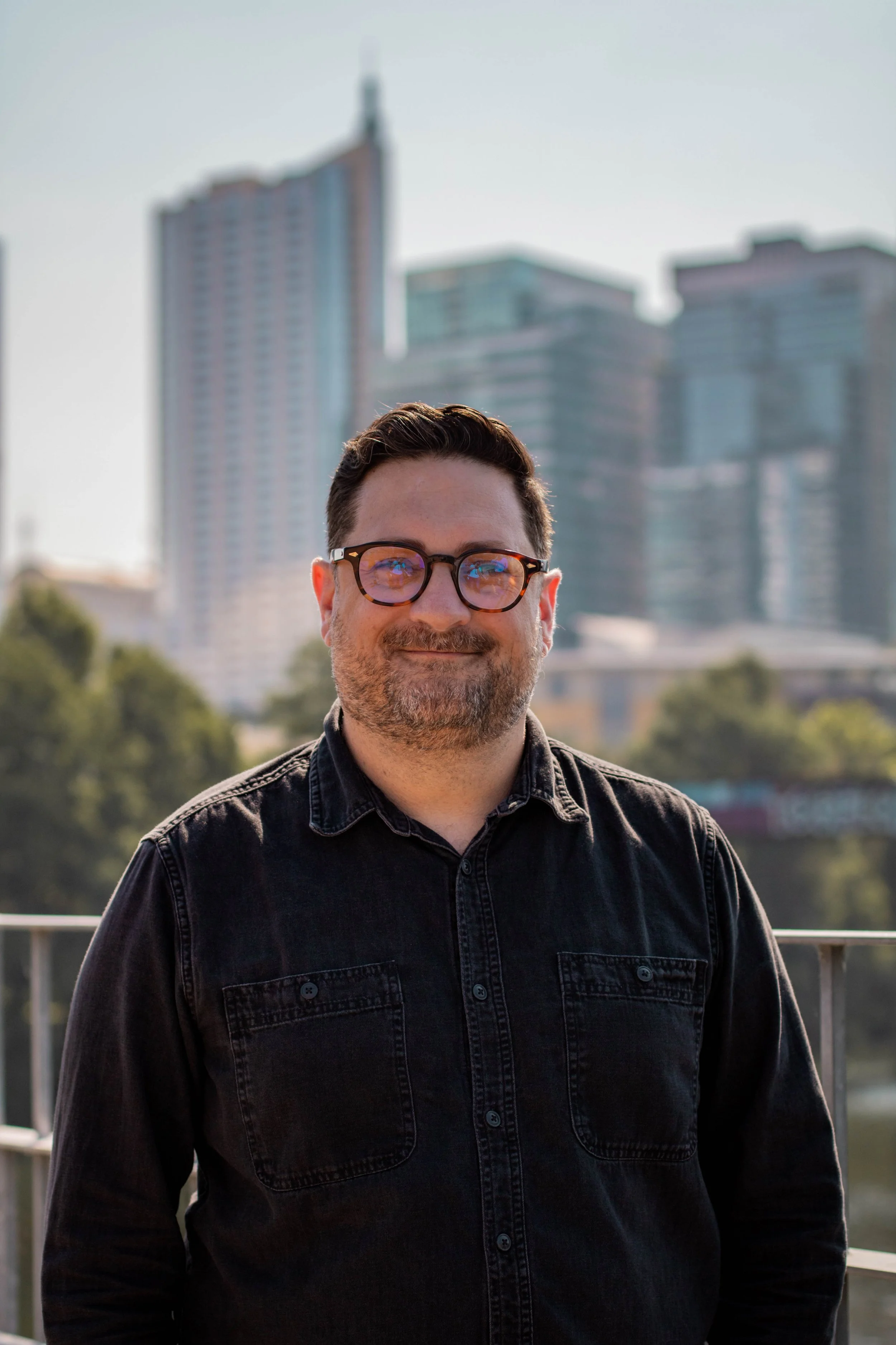 Man with glasses and beard smiling outdoors with city skyscrapers in the background.
