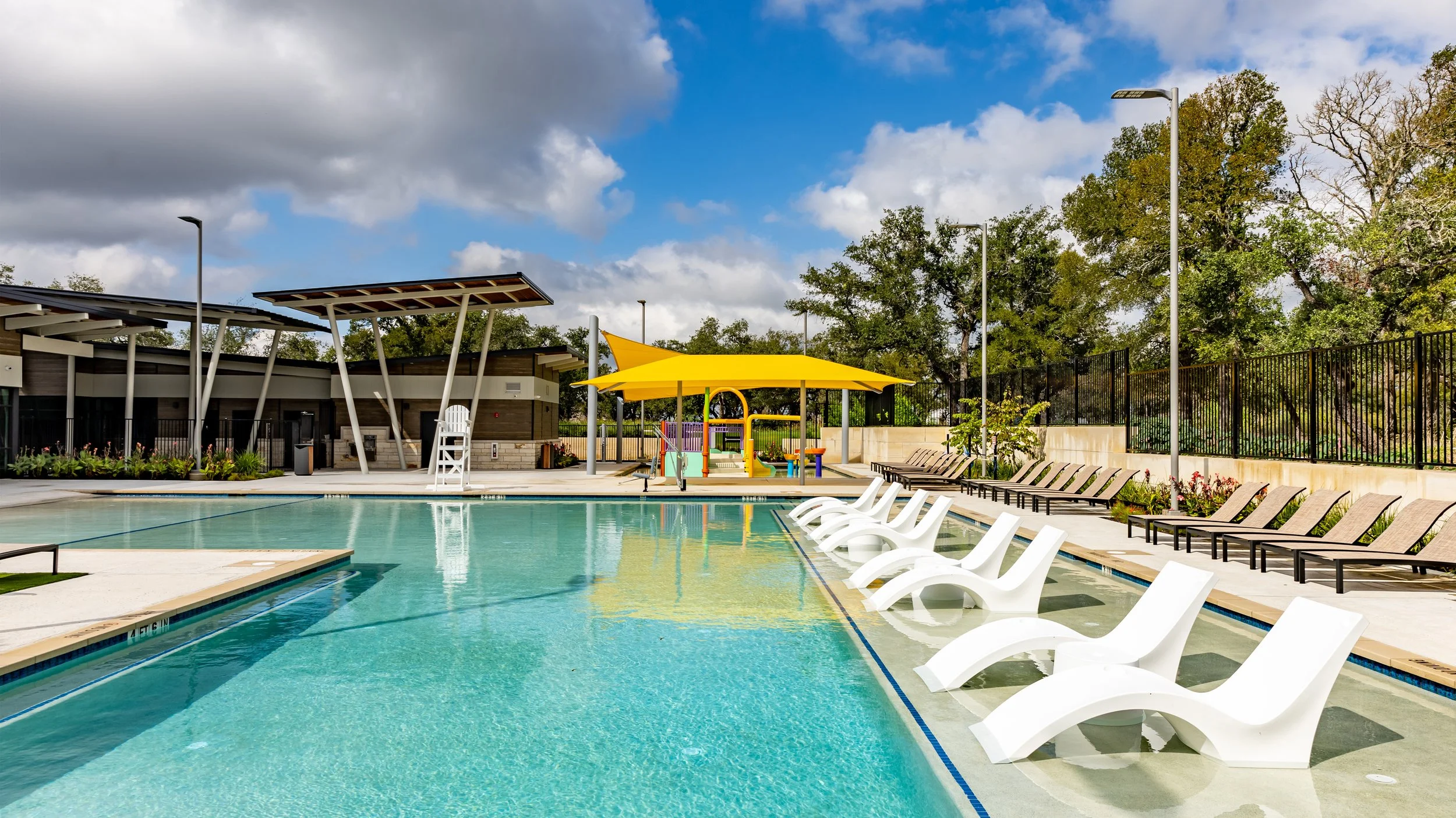Empty swimming pool with white lounge chairs aligned along the shallow end, surrounded by trees and a cloudy sky. There is a yellow shade sail and a small children’s play area in the background.