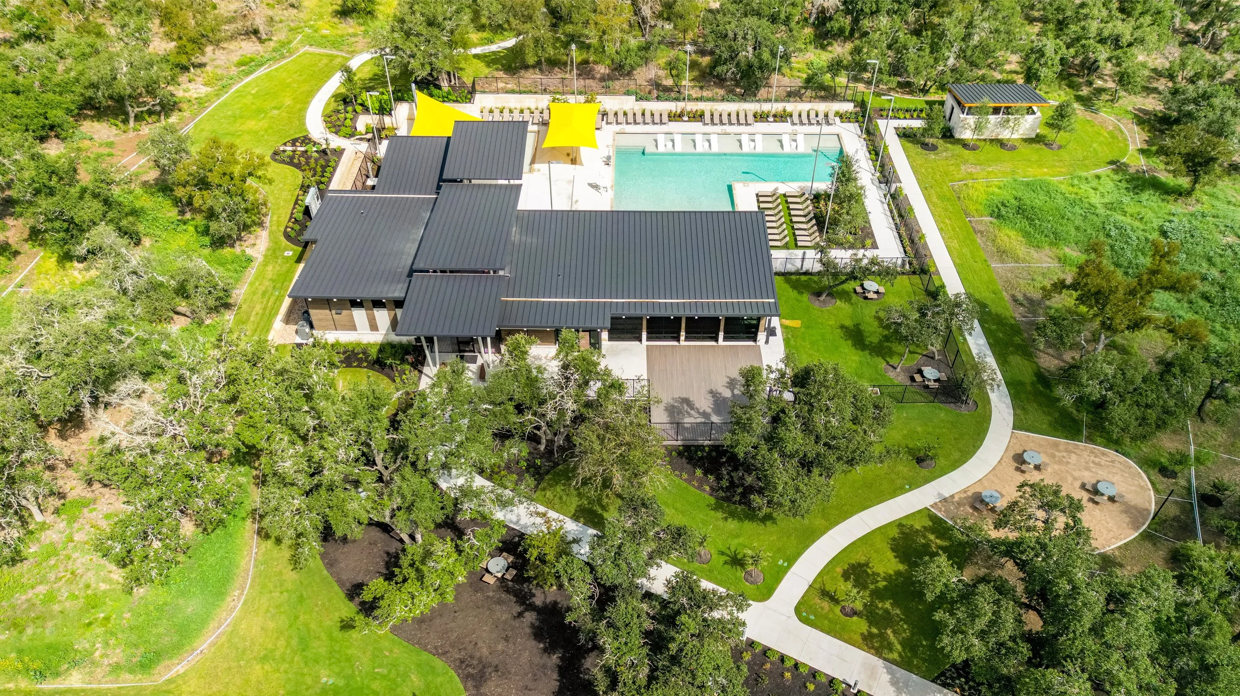 Aerial view of a modern house with a black metal roof, a swimming pool, and surrounding greenery including trees, lawns, and gardens.