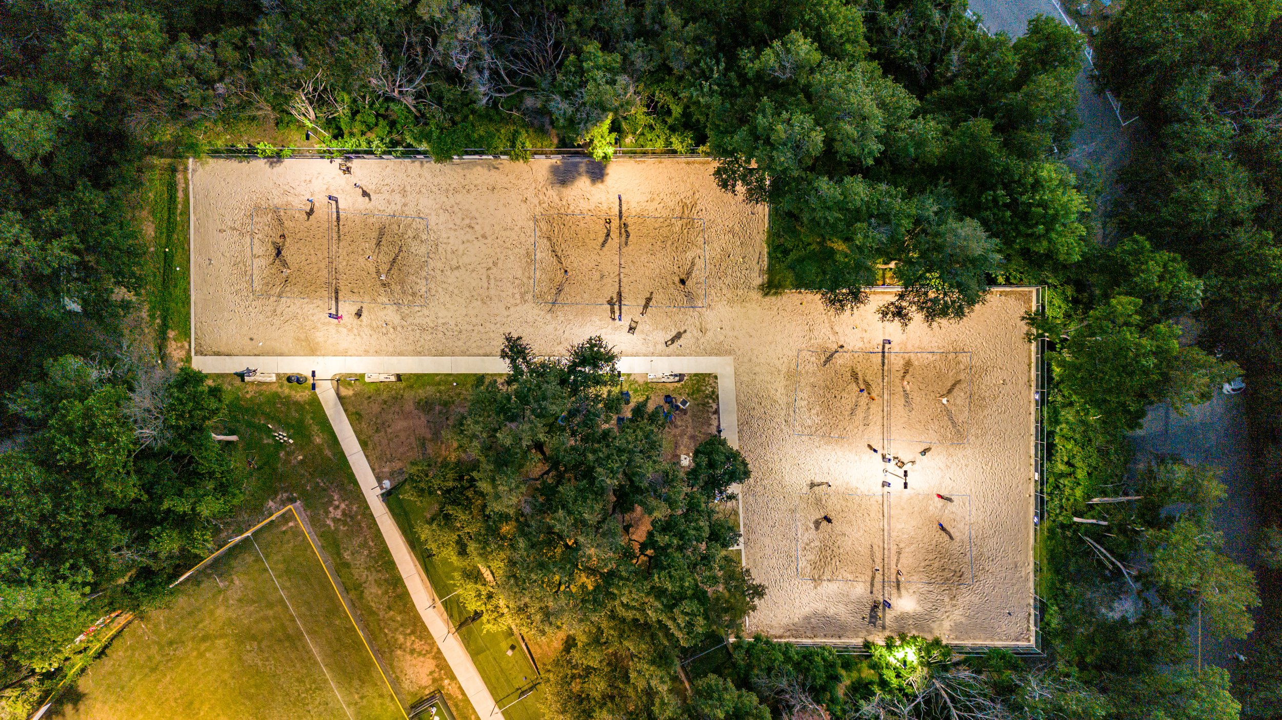 An aerial view of a sports park showing three sand volleyball courts illuminated at night, with players actively playing, surrounded by trees and a paved walkway.