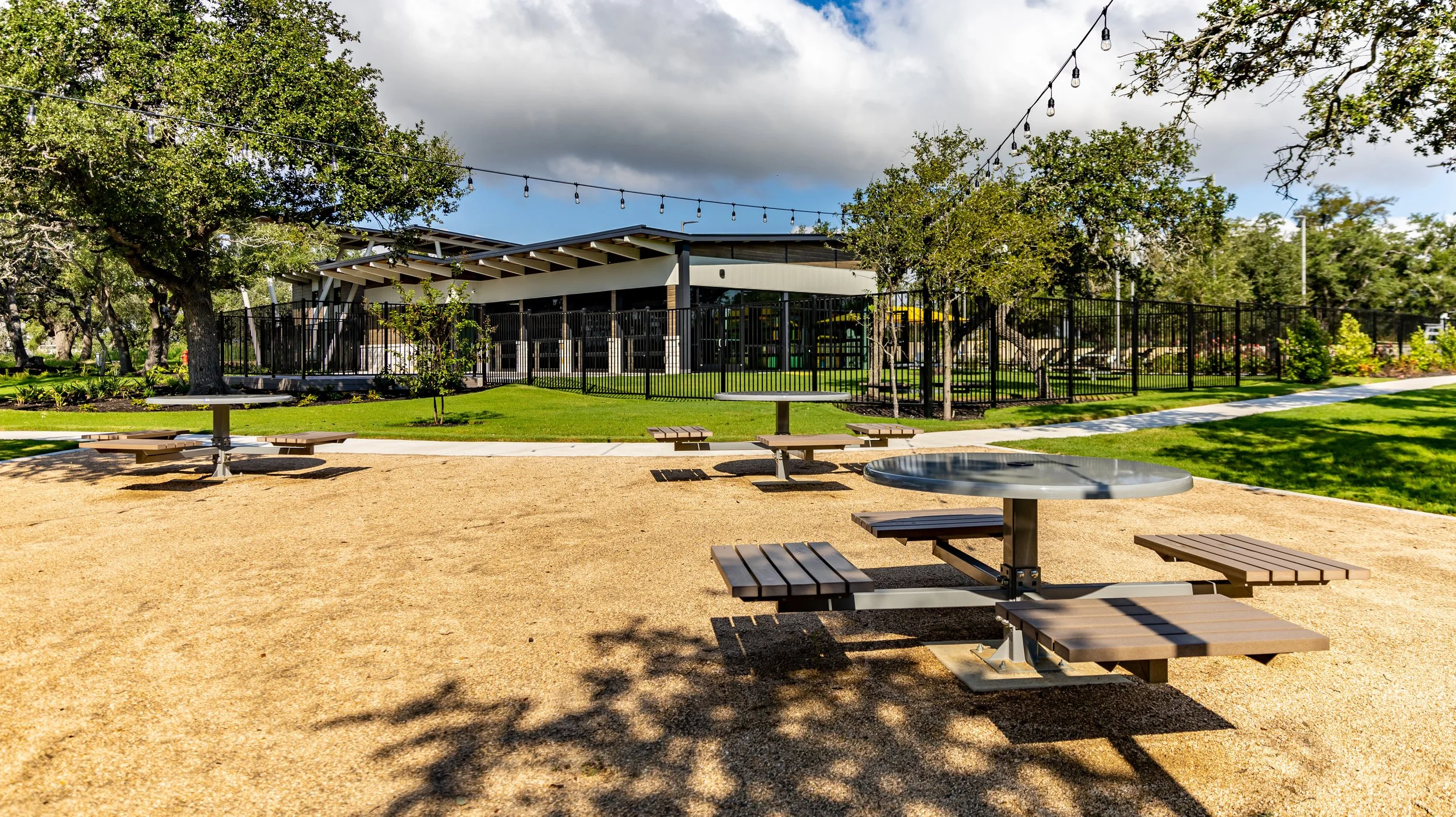 A park with a sandbox, picnic tables, trees, grassy areas, and a building with a black fence and string lights overhead.