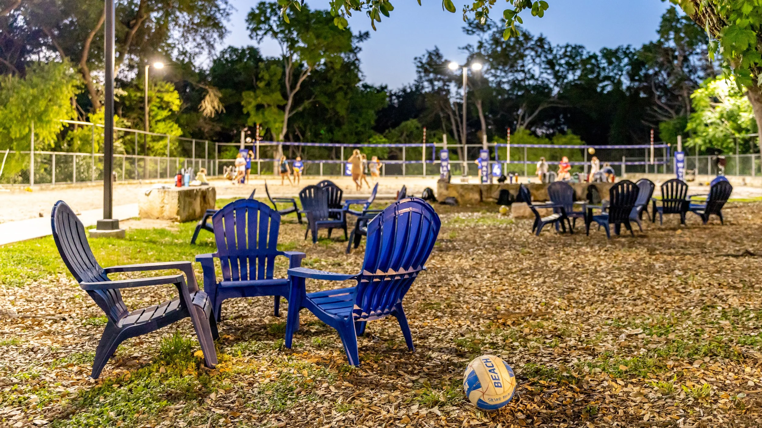 Beach volleyball court with net, outdoor seating with blue chairs, a volleyball on the ground, trees, and park lights at dusk.