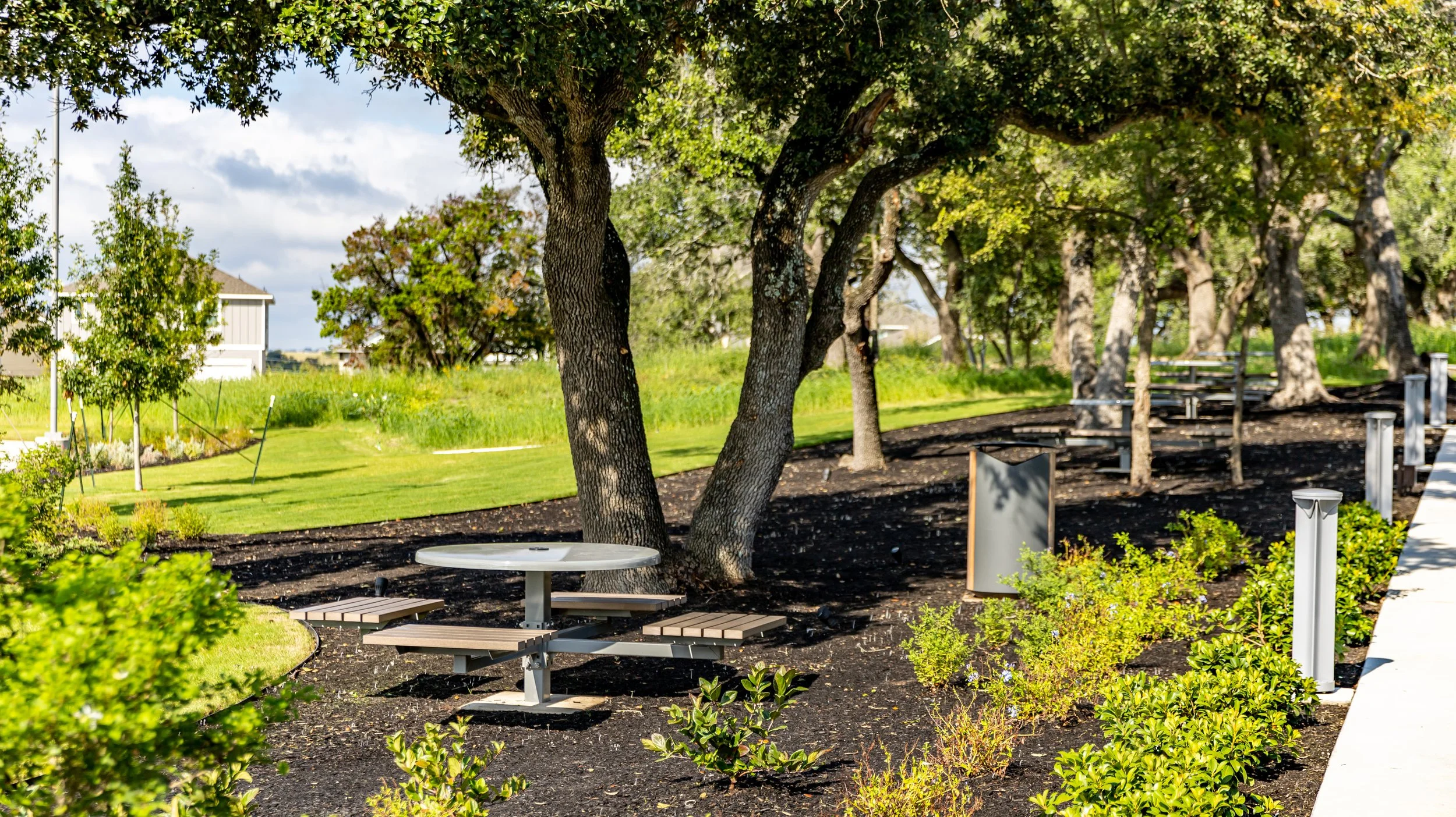 A landscaped park with large trees, bushes, and a grass area. There are picnic tables, solar-powered trash cans, and a concrete walkway.