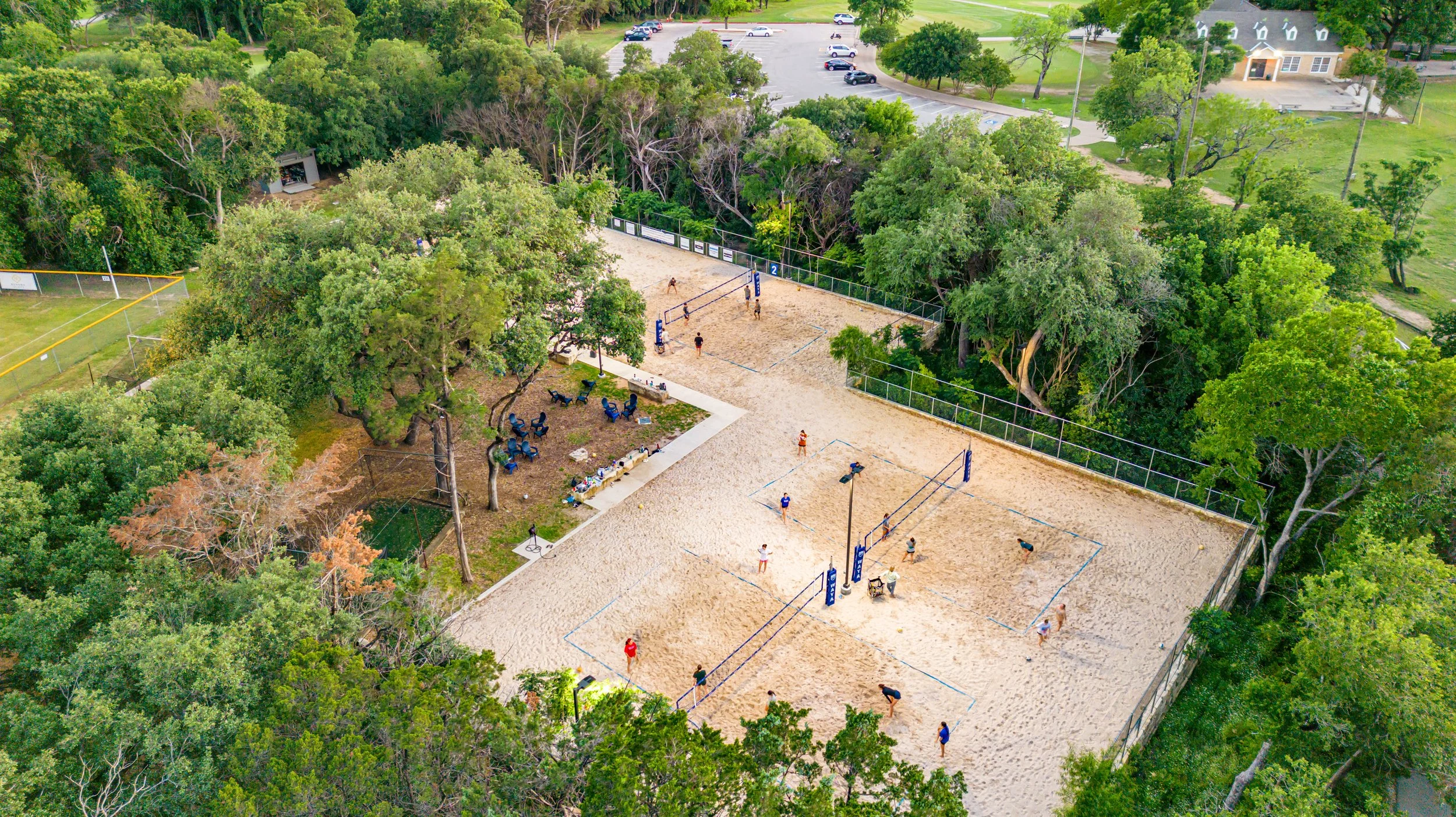 An aerial view of a sand volleyball court with six teams playing, surrounded by lush green trees, a seating area with blue chairs, and a parking lot in the background.