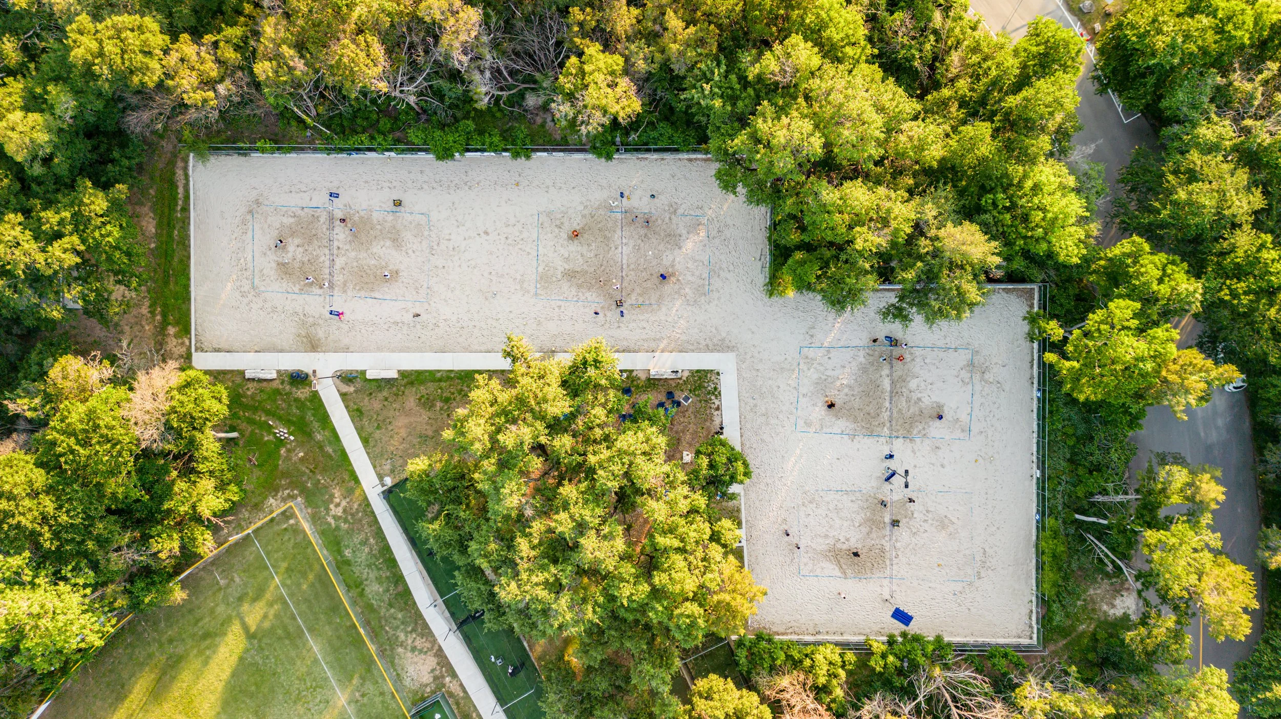 Aerial view of two sand volleyball courts surrounded by trees, with a pathway and a nearby sports field.