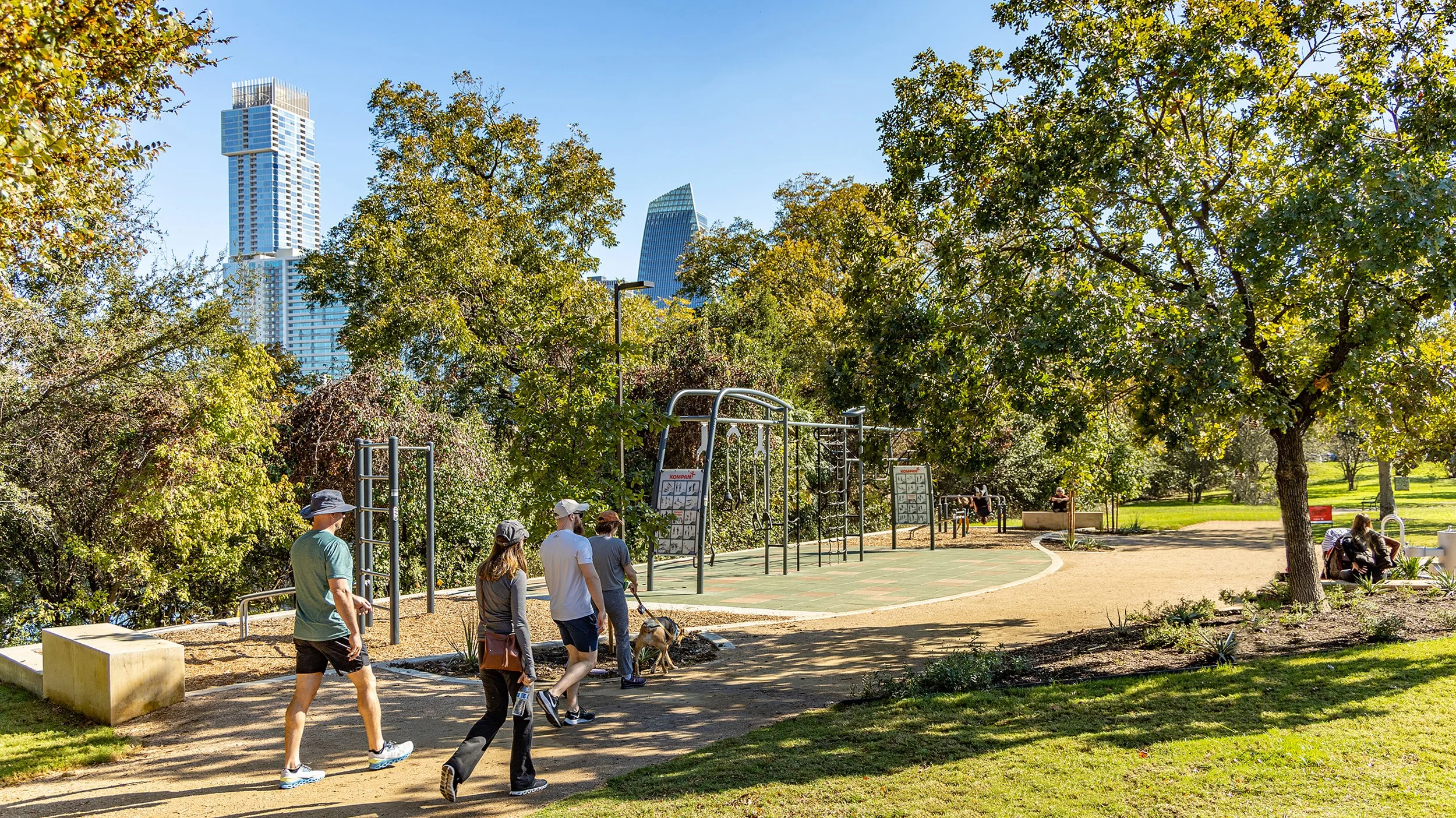 People walking in a park with trees and greenery, city skyscrapers in the background, and playground equipment visible.