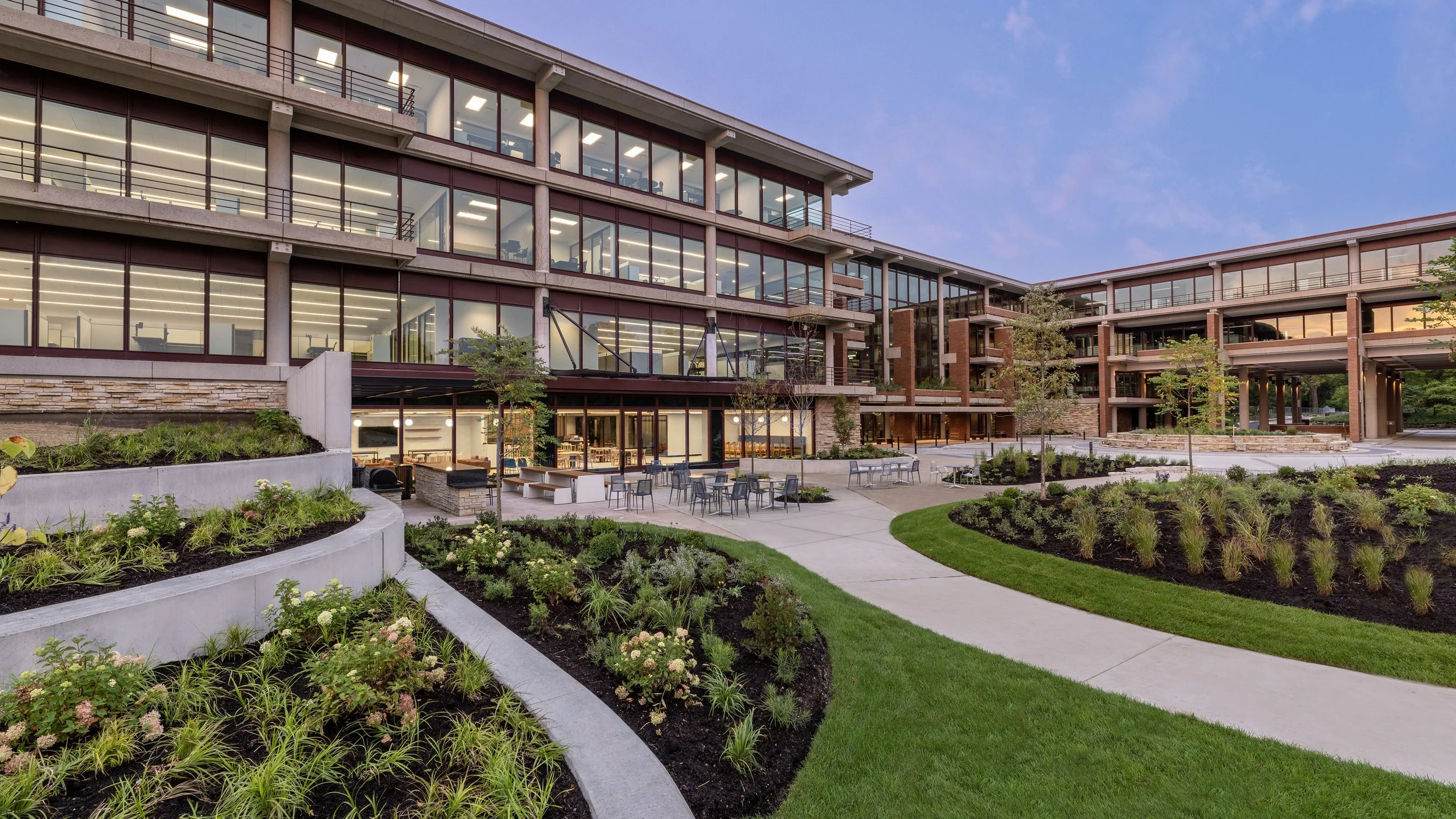 Exterior view of a modern multi-story building with large glass windows, outdoor seating area, landscaped gardens, and walking paths at dusk.