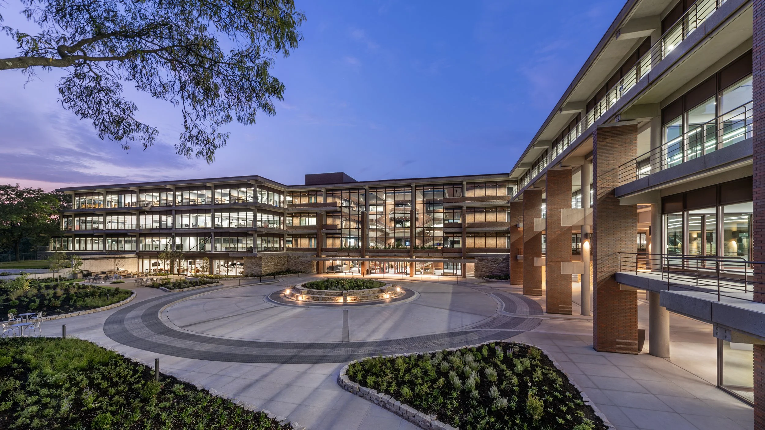 Modern multi-story building with glass walls and open walkways, illuminated and surrounded by landscaped gardens and trees at dusk.