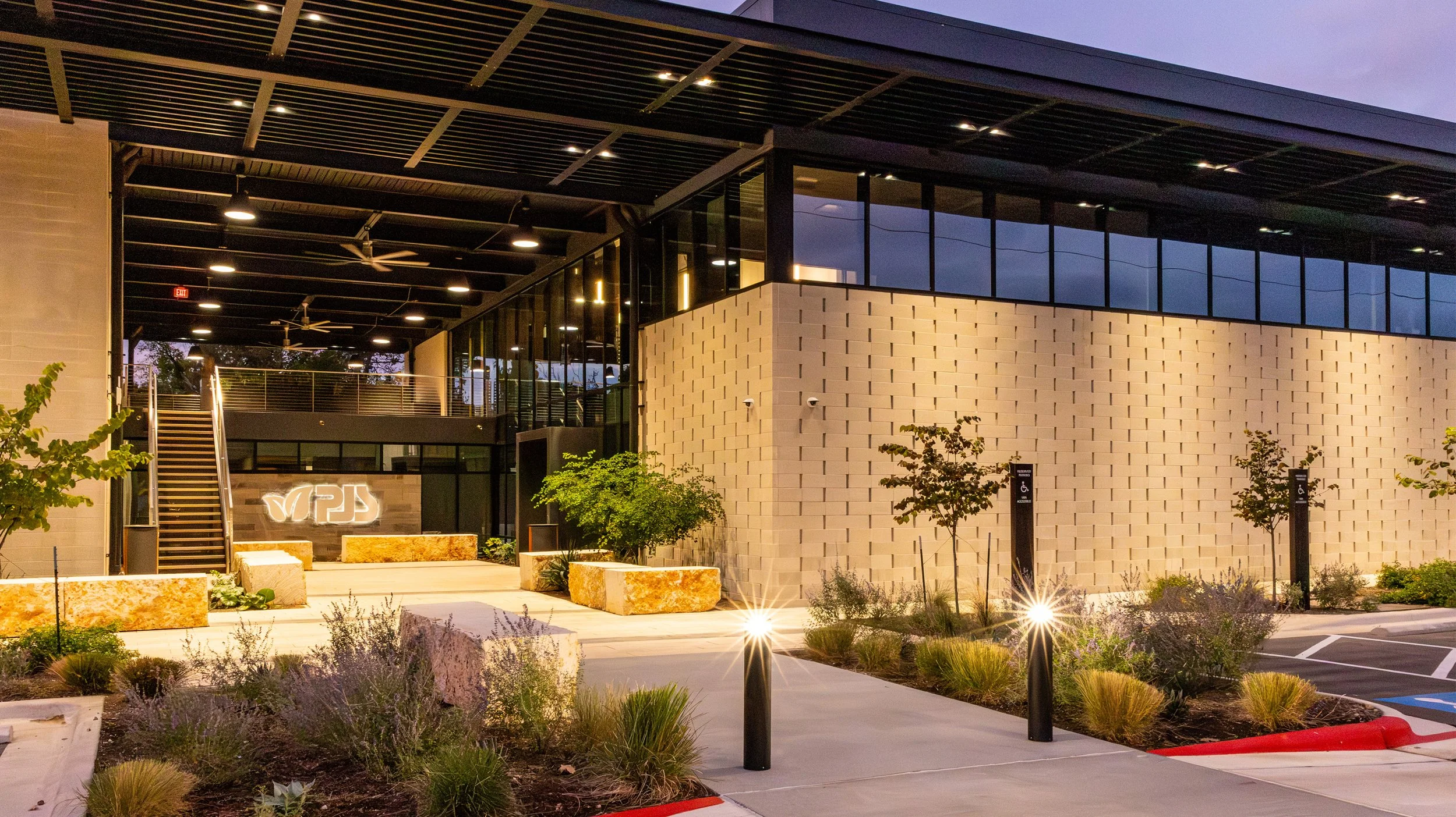 Modern commercial building with exterior landscaping, lit entrance, and designated handicap parking spaces at dusk.