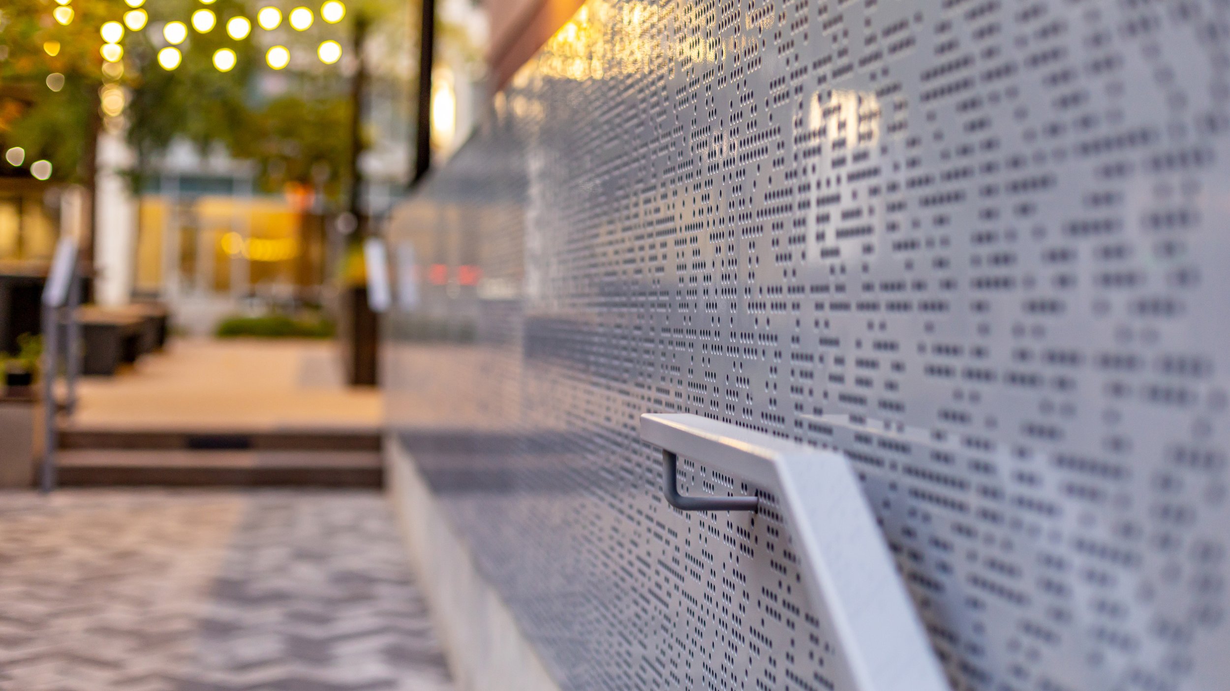 Close-up of a memorial wall with engraved names, with a blurred outdoor background featuring trees and lights.