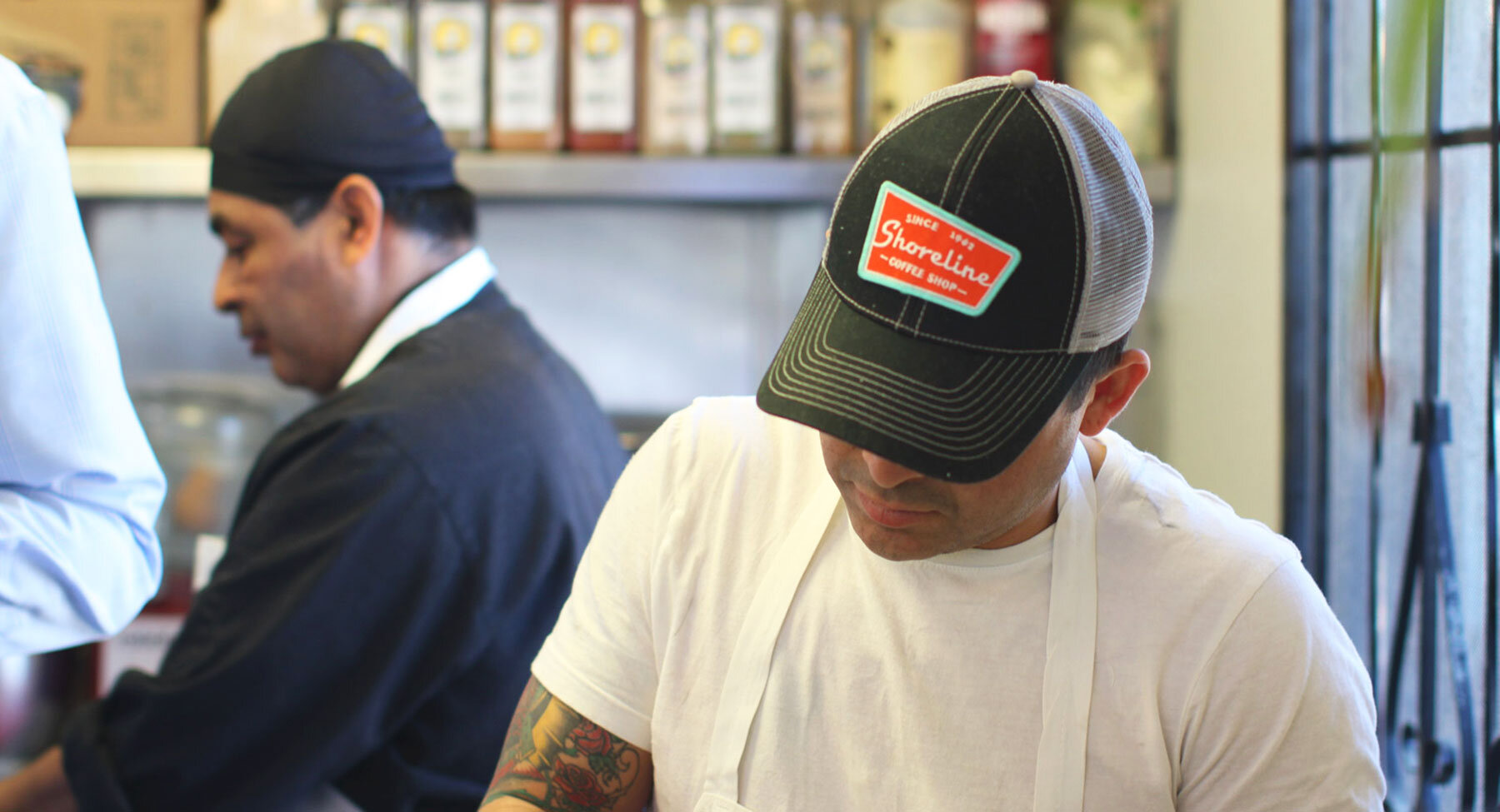 A man wearing a black shirt and a second man wearing a white shirt with a Shoreline Coffee Shop branded baseball hat