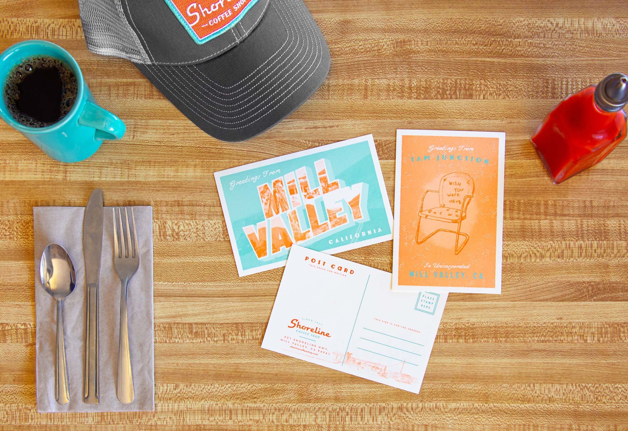 Three postcards with Shoreline Coffee Shop branding on a wooden surface next to silverware, a cup of coffee and a hat