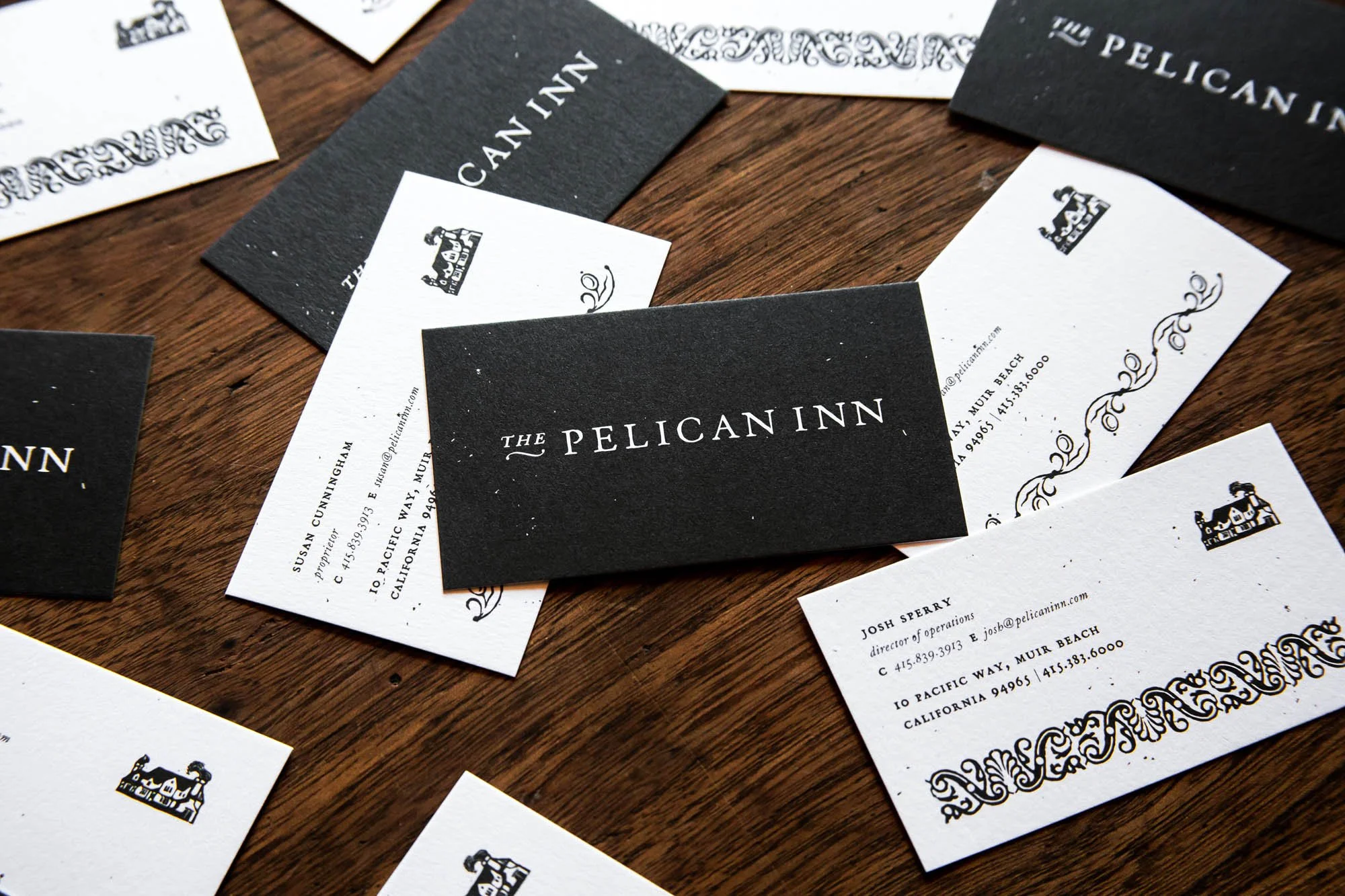 Business cards for The Pelican Inn scattered on a wooden surface, featuring black and white designs with ornate motifs and contact information.