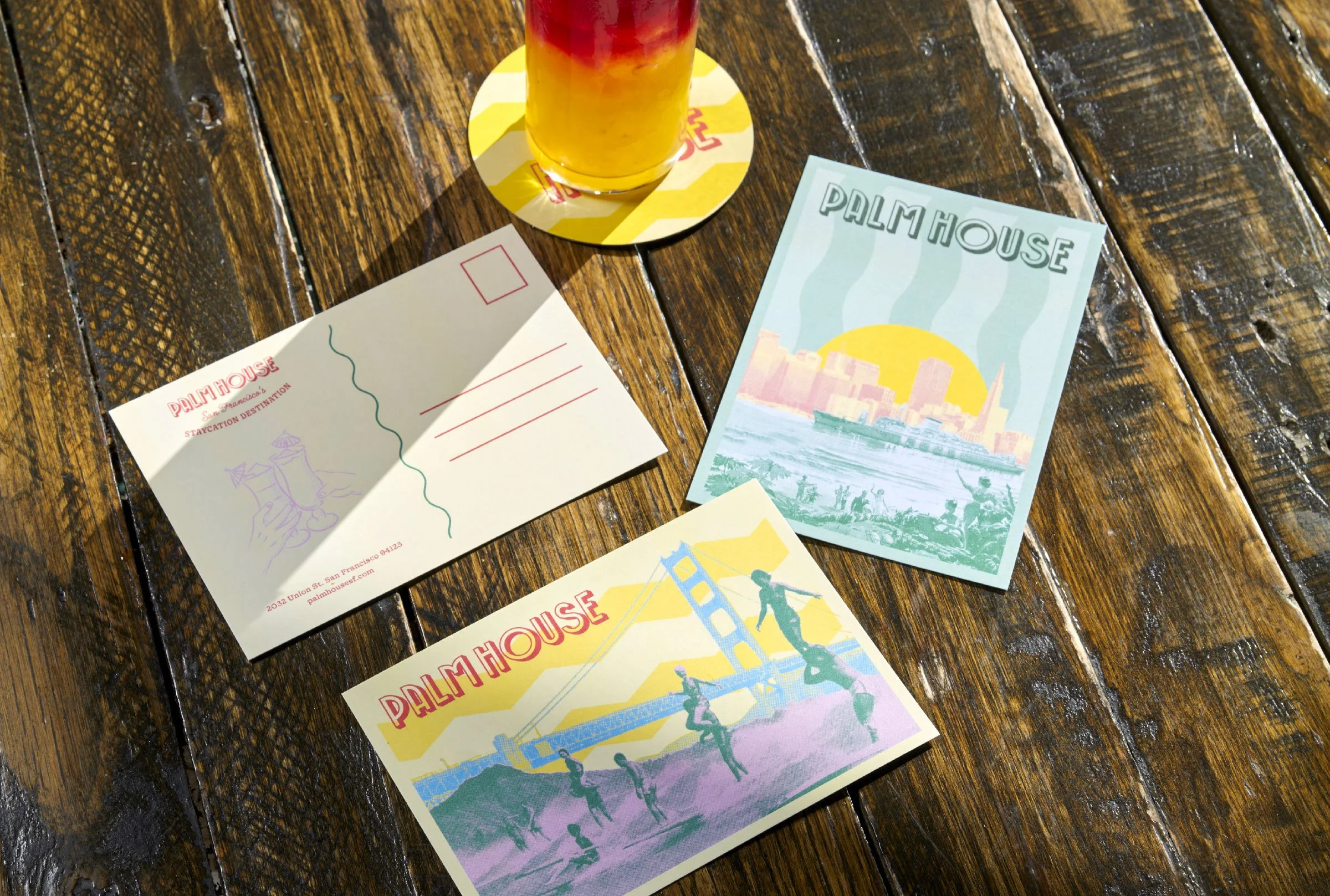 Postcards from Palm House featuring cityscape, bridge, and sunset, with a drink on a wooden table.