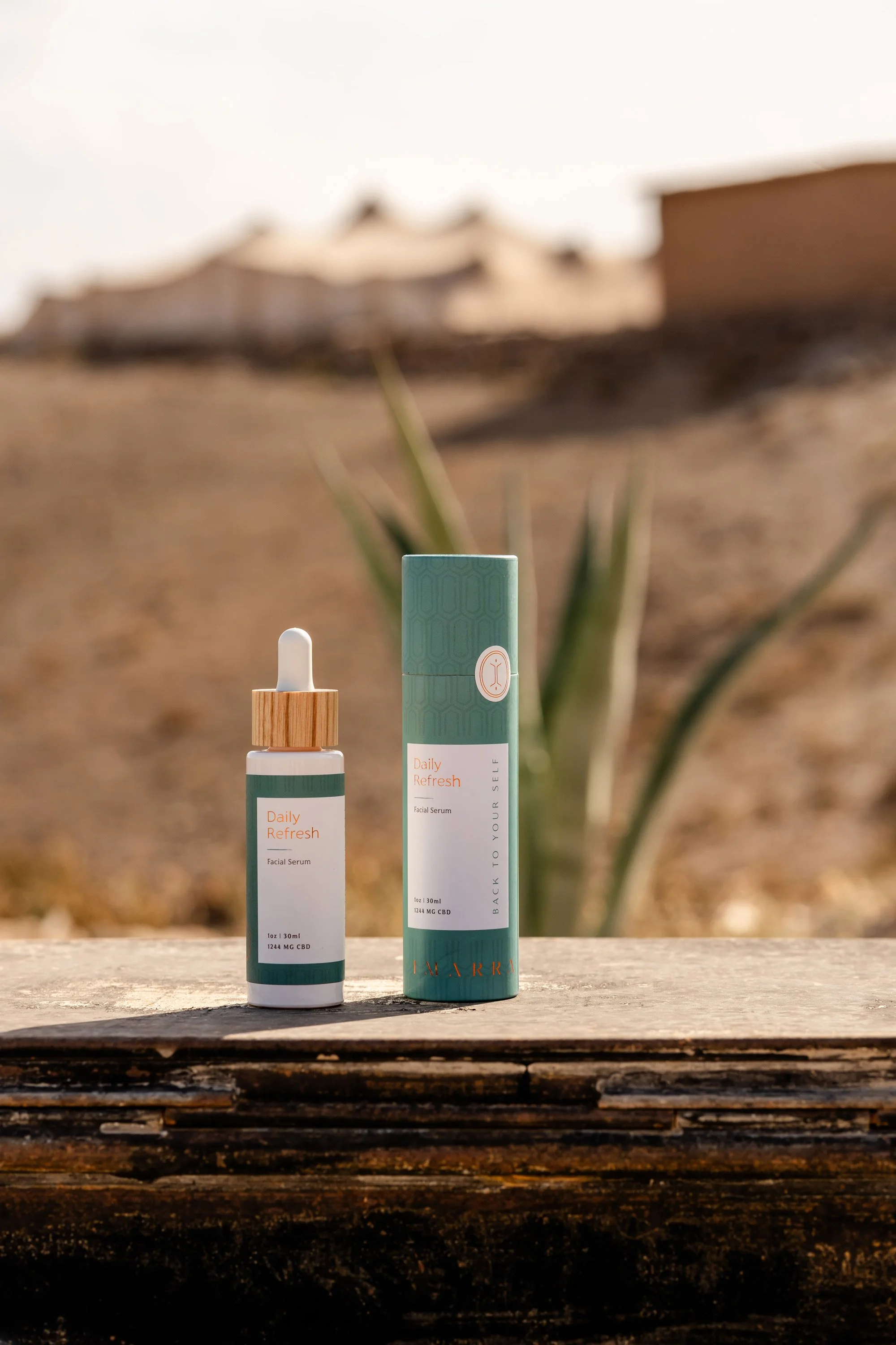 A facial serum bottle and matching box labeled 'Daily Refresh' are placed on a wooden surface outdoors, with a blurred desert landscape and plant in the background.