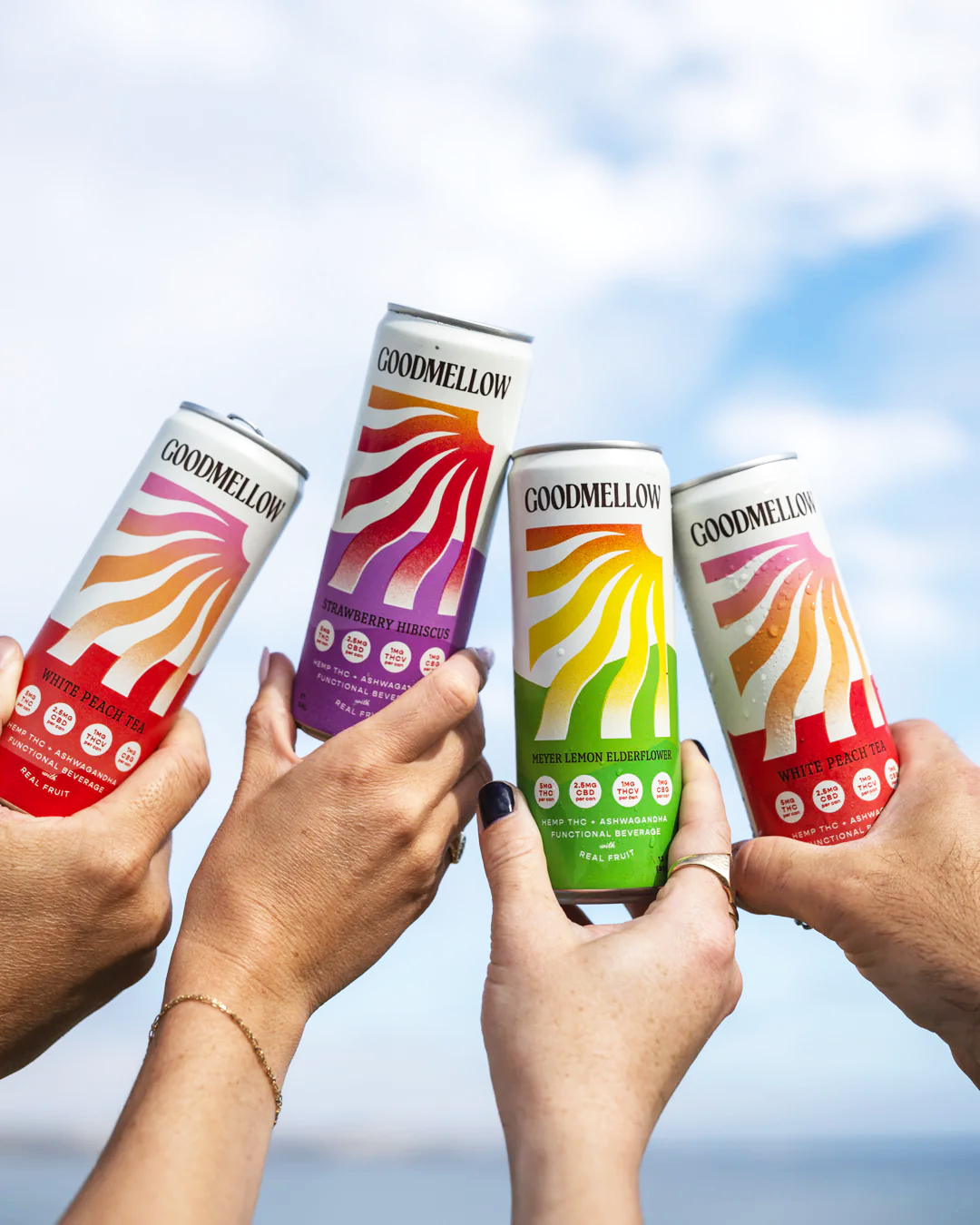 Four hands holding cans of Goodmellow functional beverage with colorful labels and a partly cloudy sky in the background.