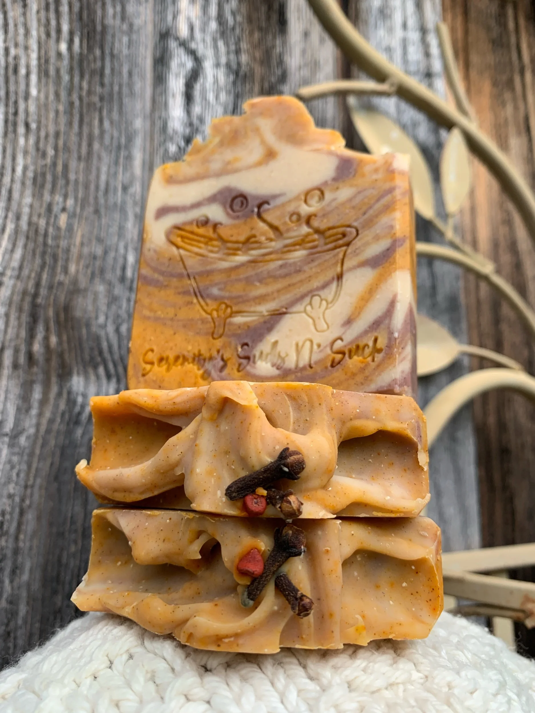 A hand holding beautiful handcrafted goat milk soaps with blue skies in the background and greenery.