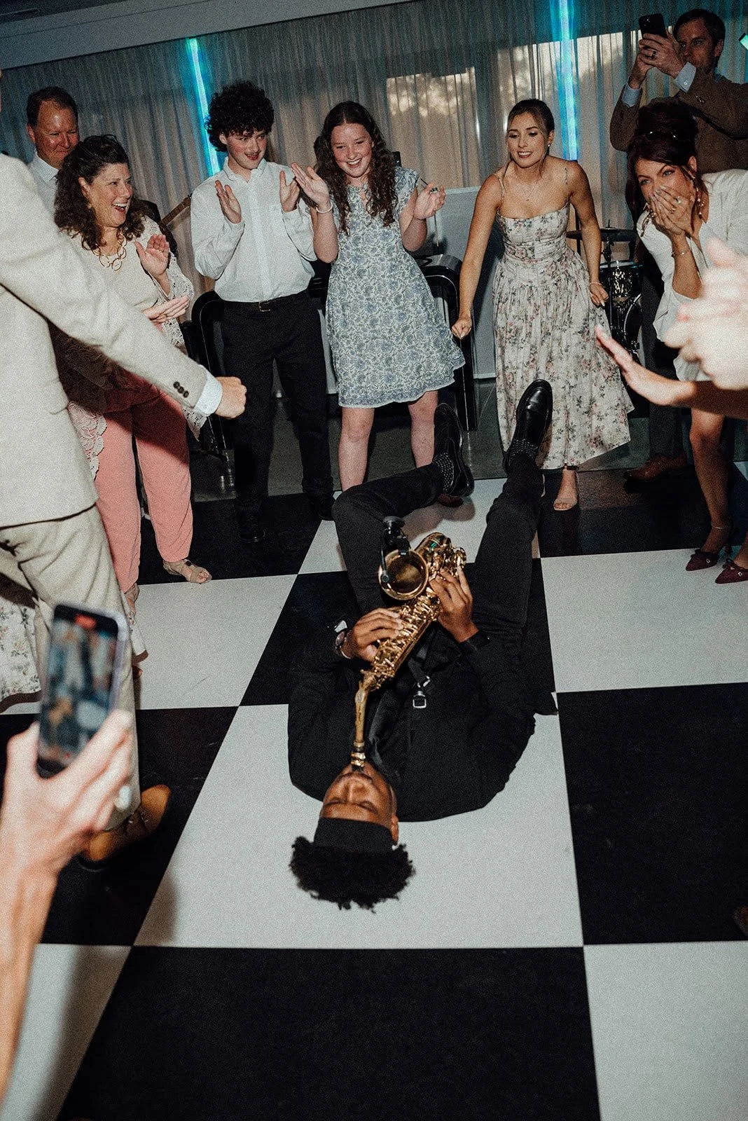 A musician lying on the black and white checkered floor, playing a saxophone, surrounded by a group of smiling and clapping people at a celebration or party.