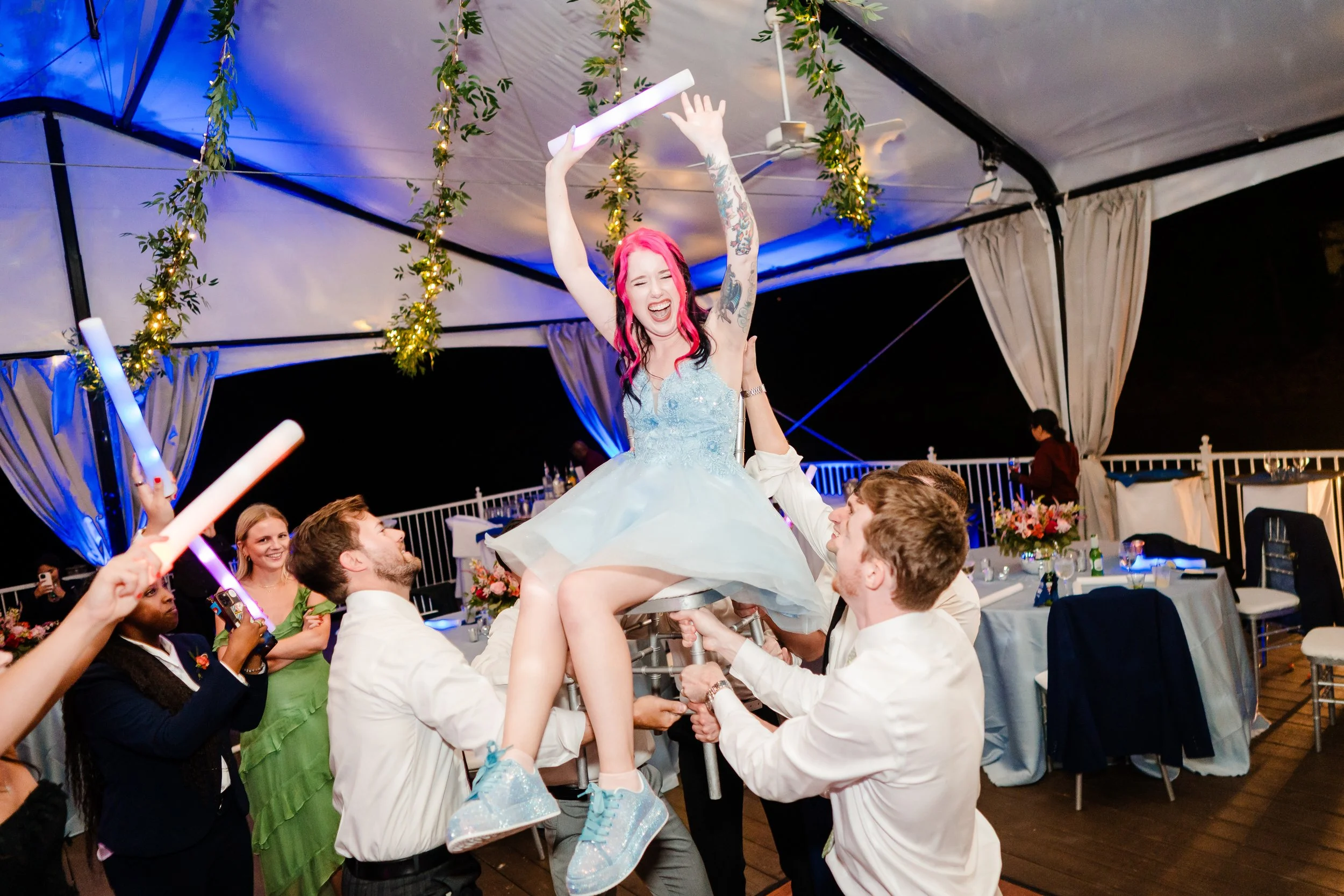A woman with pink hair and tattoos on her arm, wearing a light blue dress, is being lifted on a chair by multiple people during a celebration at a decorated venue with string lights and floral arrangements.