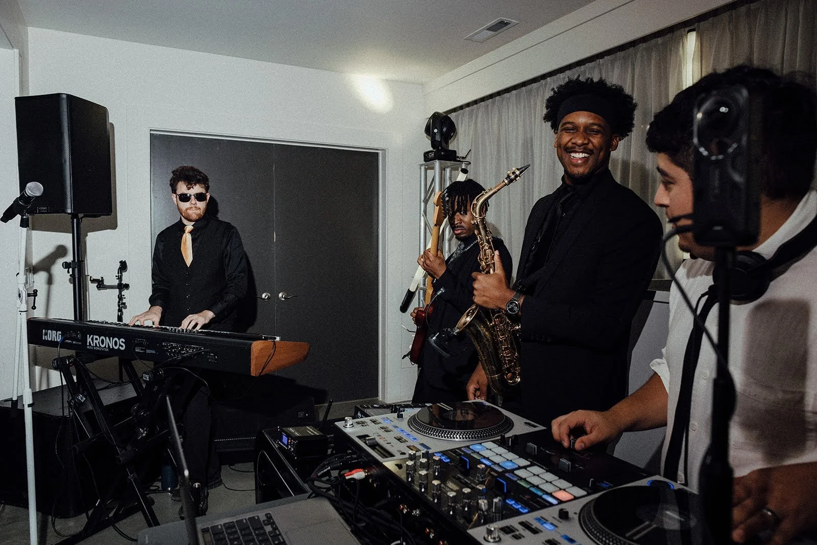 A group of four musicians performing indoors, with a keyboard, saxophone, DJ controller, and microphone, in a room with closed curtains and a door behind them.