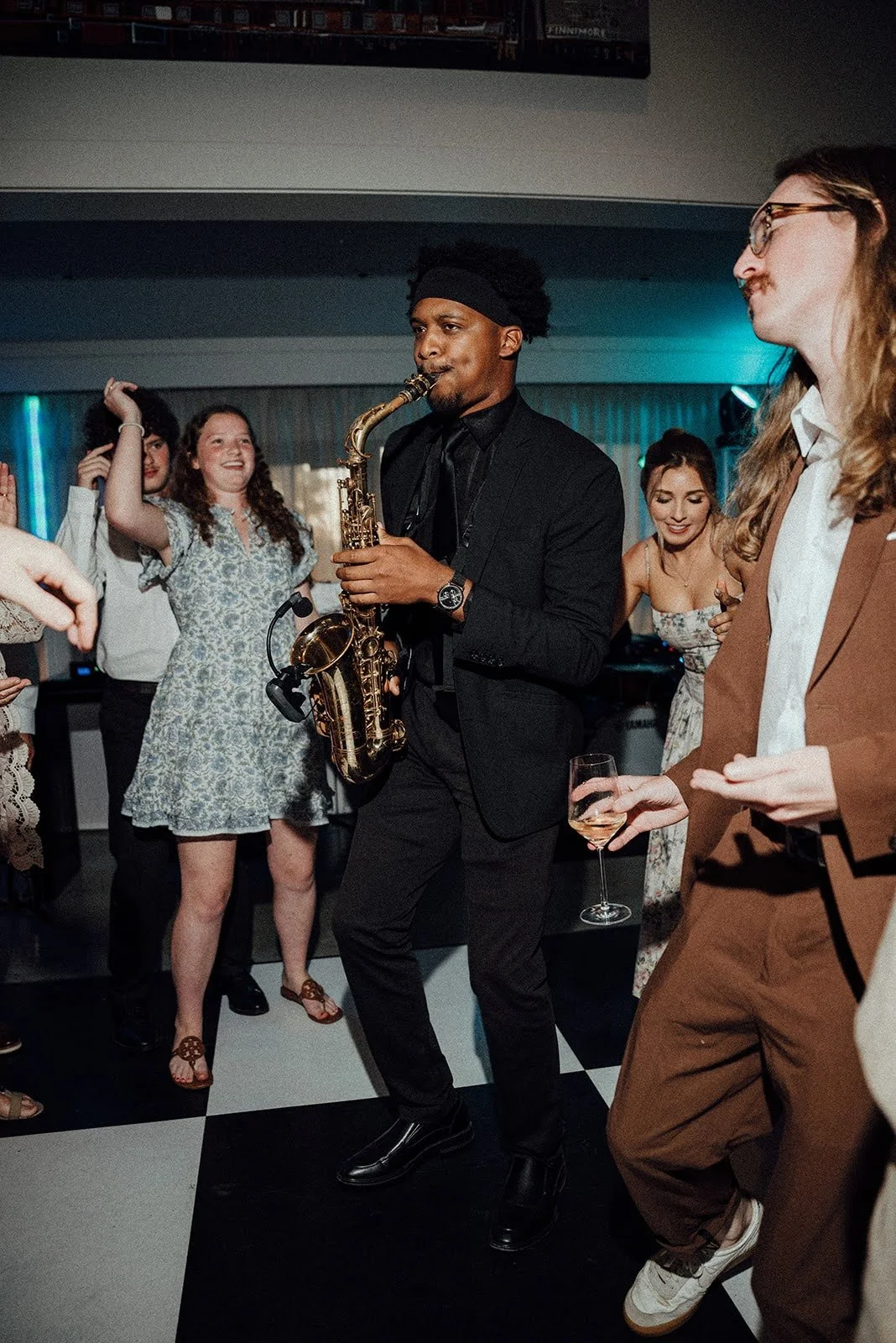 A man in a black suit and headband playing a saxophone while dancing at a party, surrounded by smiling women holding drinks and enjoying the music.