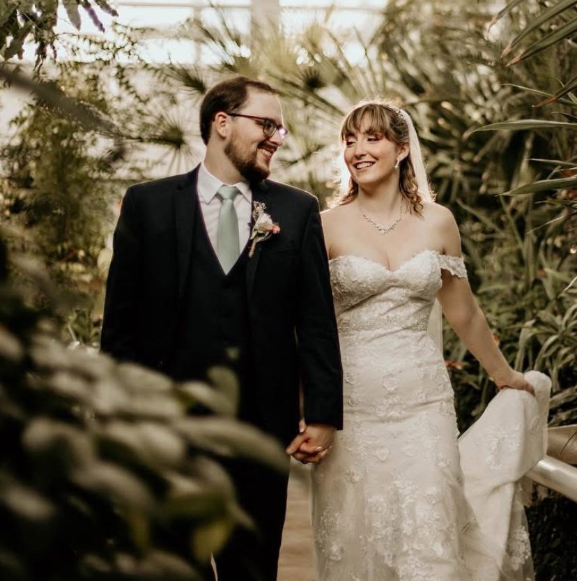 A bride and groom walking hand in hand in a lush, tropical greenhouse, both smiling and looking at each other.
