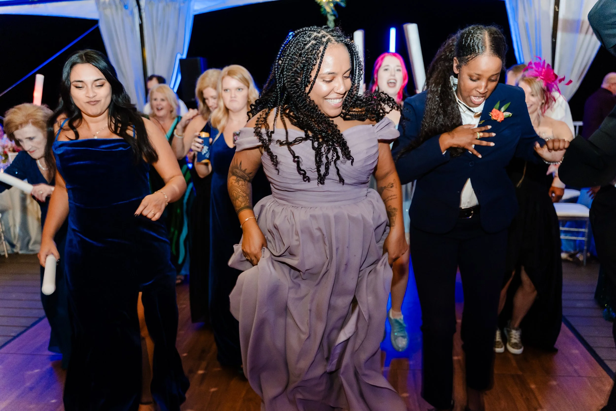 Group of women dancing and having fun at a party or celebration, under colorful lighting, with some women smiling and wearing formal dresses.