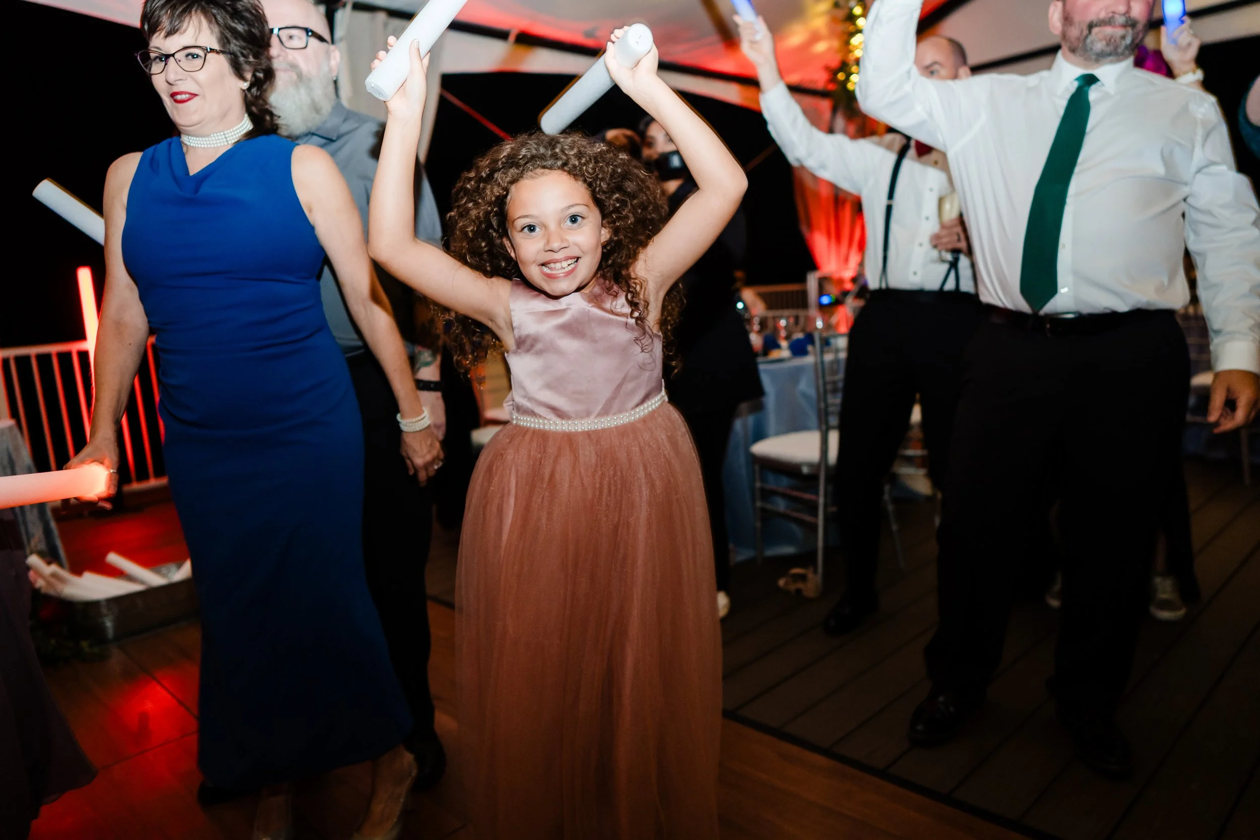A group of people dancing at a party, with a young girl in a pink dress smiling and holding glow sticks in the foreground.